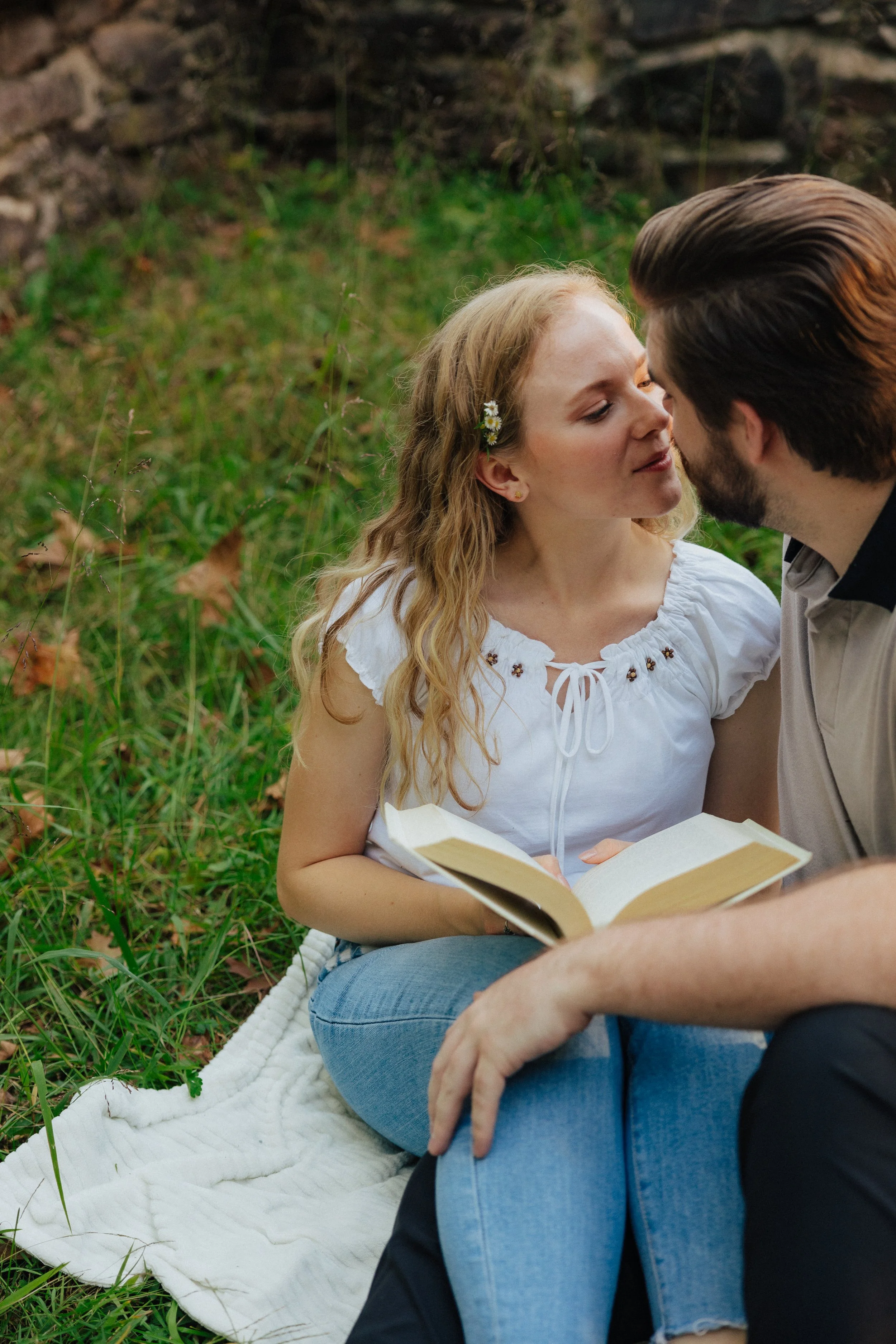 A young woman with long, curly blonde hair and a flower hair accessory, wearing a white embroidered blouse, and a man with dark hair and beard, wearing a beige shirt, are sitting on a white blanket outdoors in a grassy area. They are close together, 