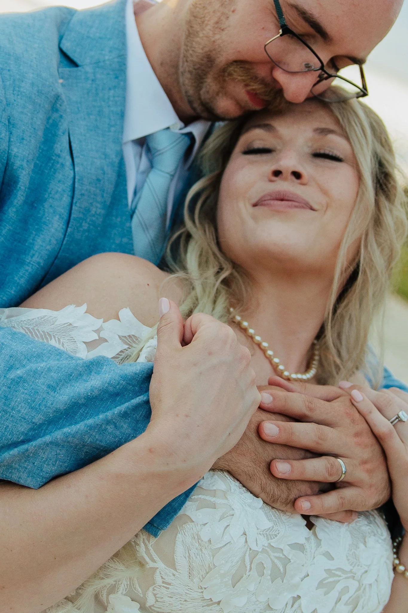 A man in a blue suit and glasses passionately kisses a woman in a wedding dress, holding her close with her eyes closed, wearing a pearl necklace and lapel pin.