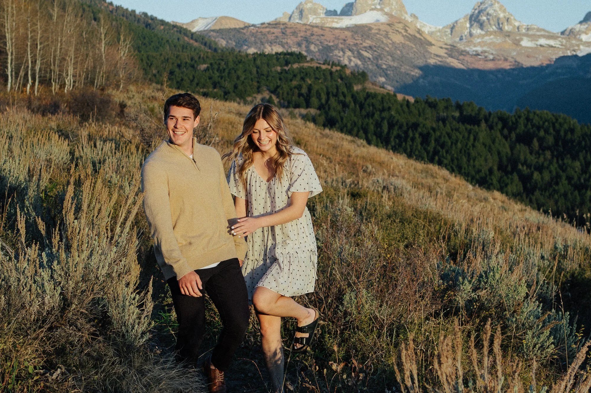 A smiling young man and woman stand together outdoors in a natural landscape with mountains and forest in the background. The woman is wearing a white polka dot dress and black sandals, and the man is in a beige sweater and black pants. They appear h