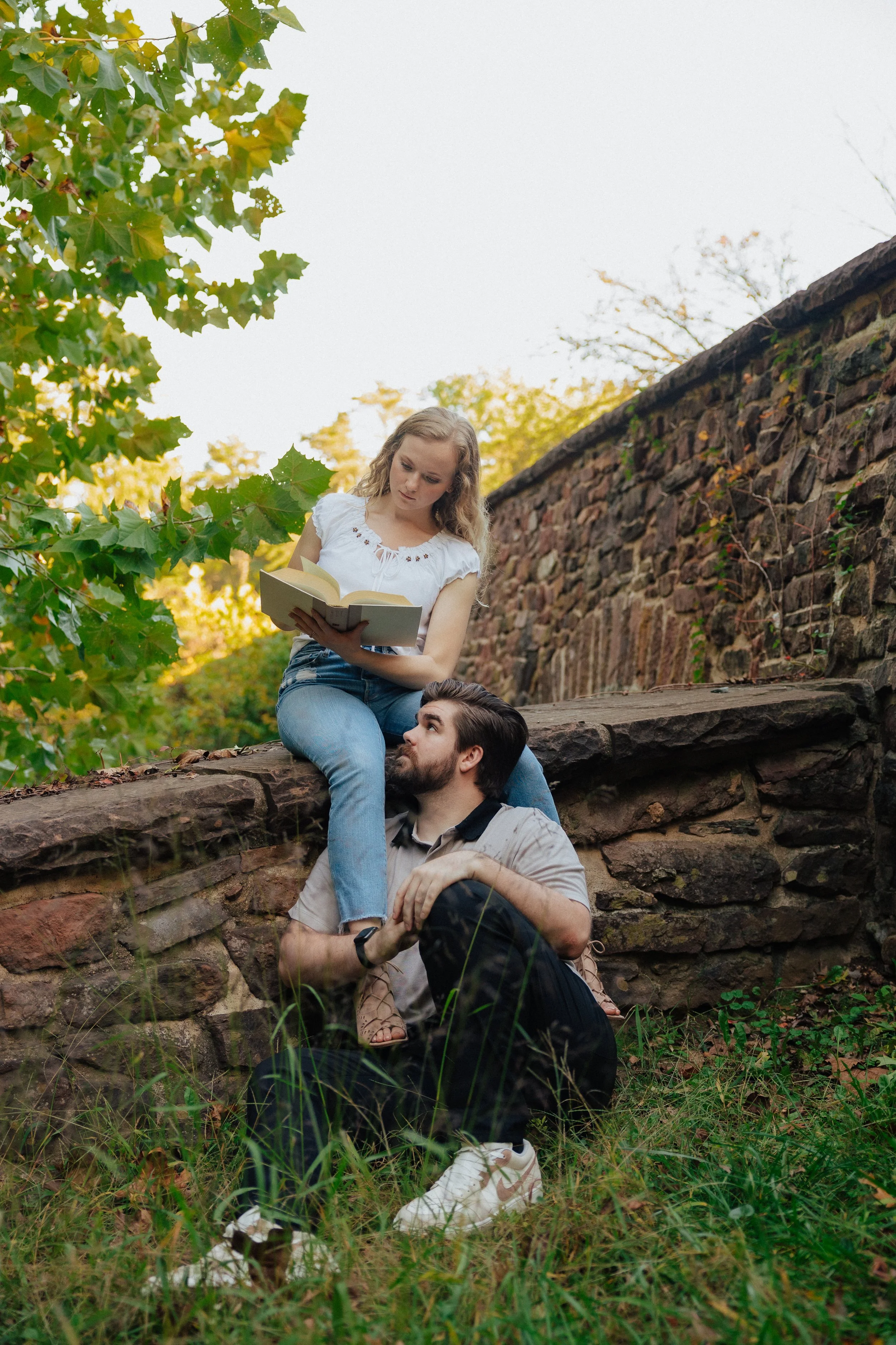 A young woman with blonde curly hair and a white top is sitting on a stone wall, reading a book. Below her, a young man with dark hair, a beard, and a beige shirt is sitting on the grass, looking up at her. They are outdoors near greenery and a stone