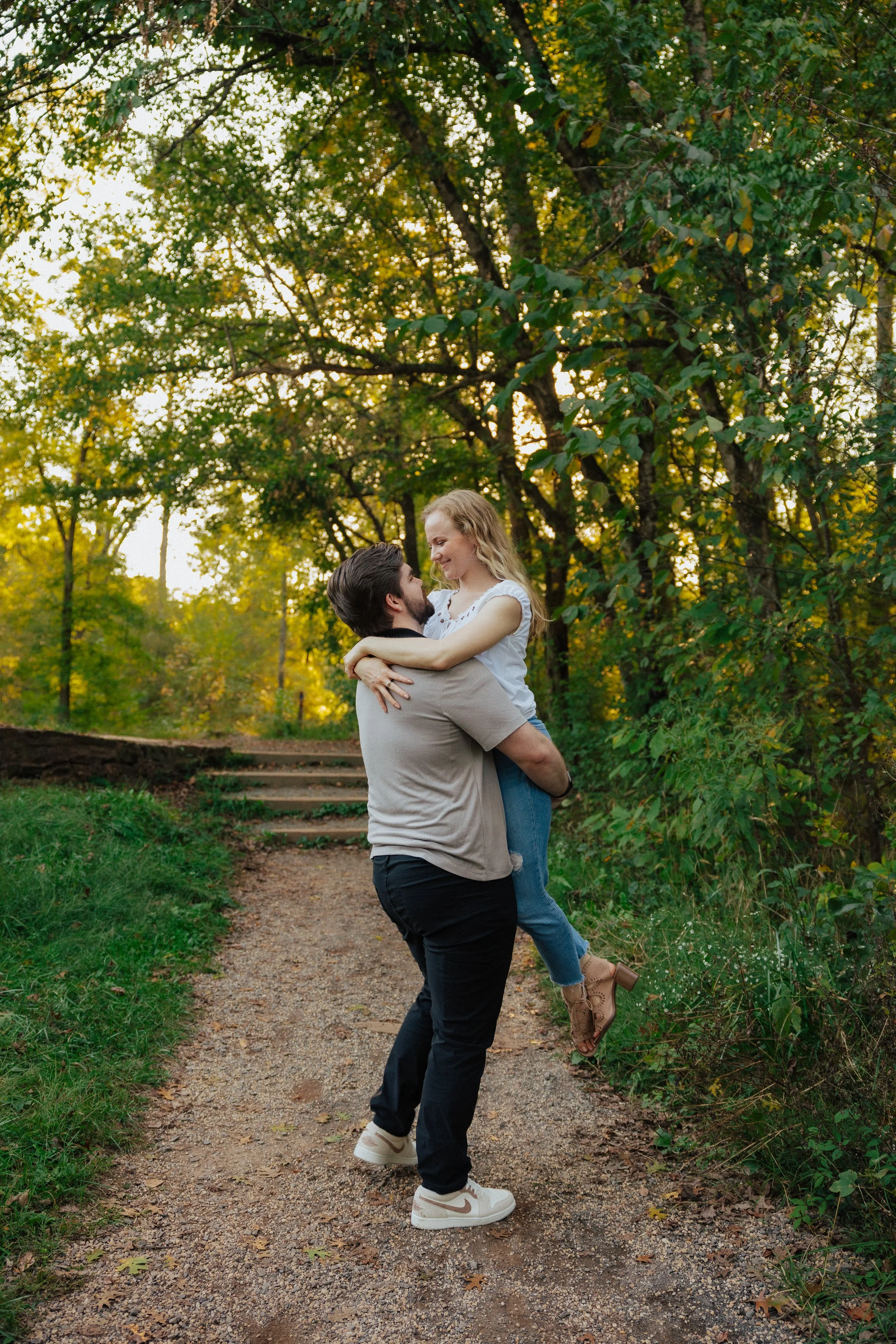 A man is holding a woman in his arms on a dirt path in a green park during fall. The woman is smiling and looking at the man, and the man is looking at her. There are trees and foliage around them with yellow and green leaves.