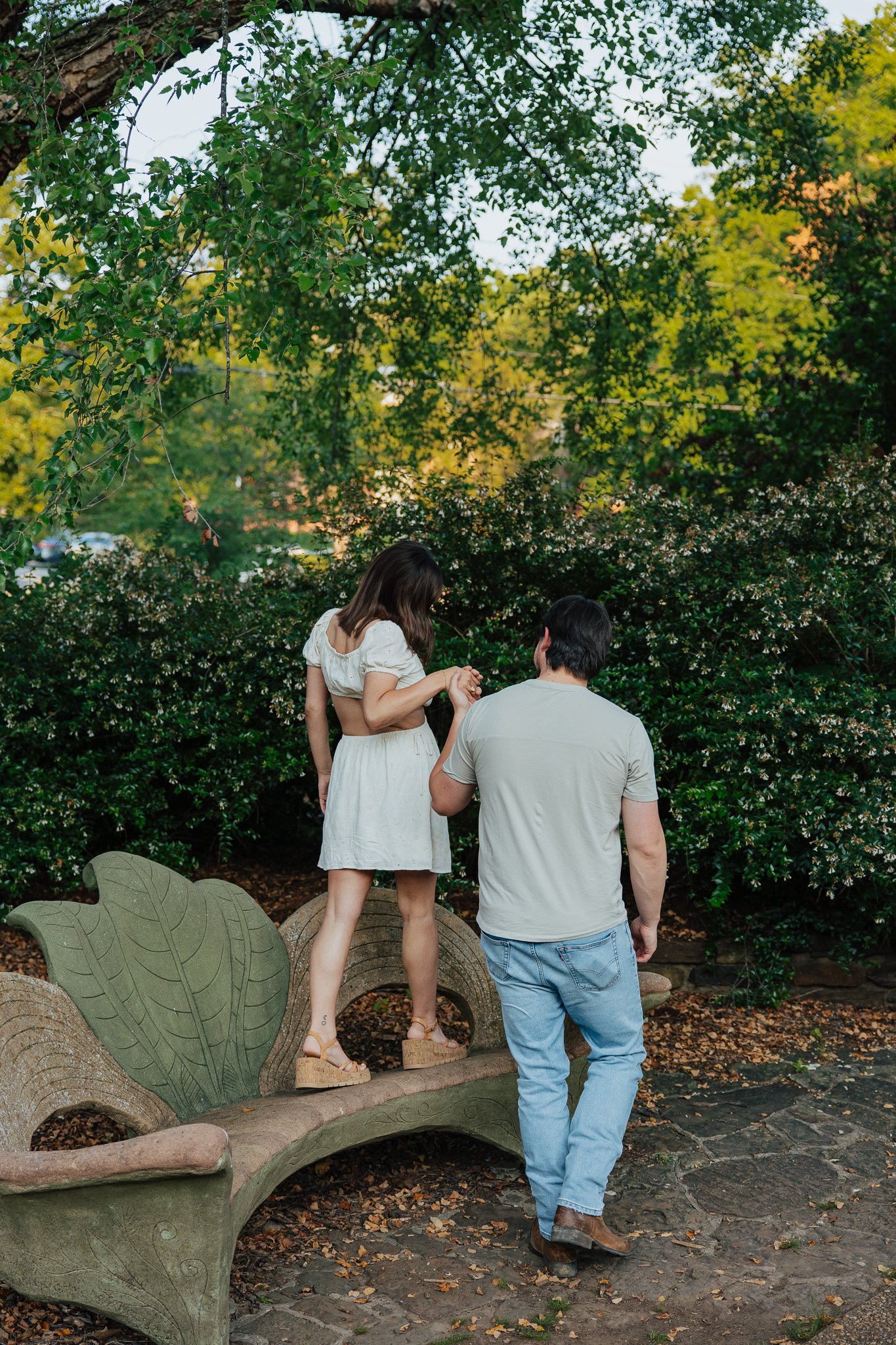 Two women and one man standing outdoors on a stone arch bridge with decorative leaf motifs, surrounded by lush green foliage, trees, and autumn leaves on the ground. The woman with brown hair is wearing a white crop top and skirt, and tan platform sh