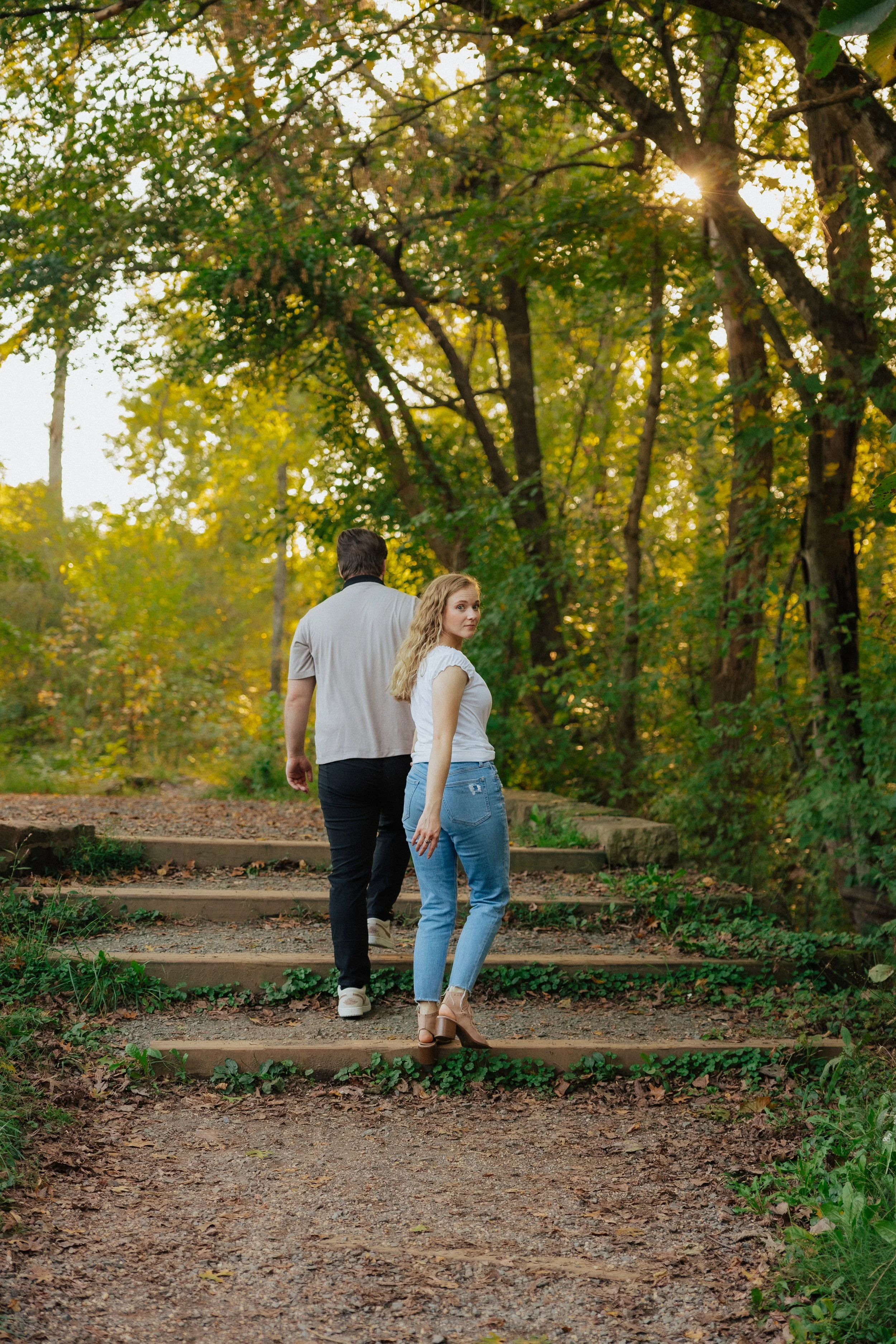 A man and woman walking up wooden steps in a forested park during daytime, with sunlight filtering through green and yellow autumn leaves.