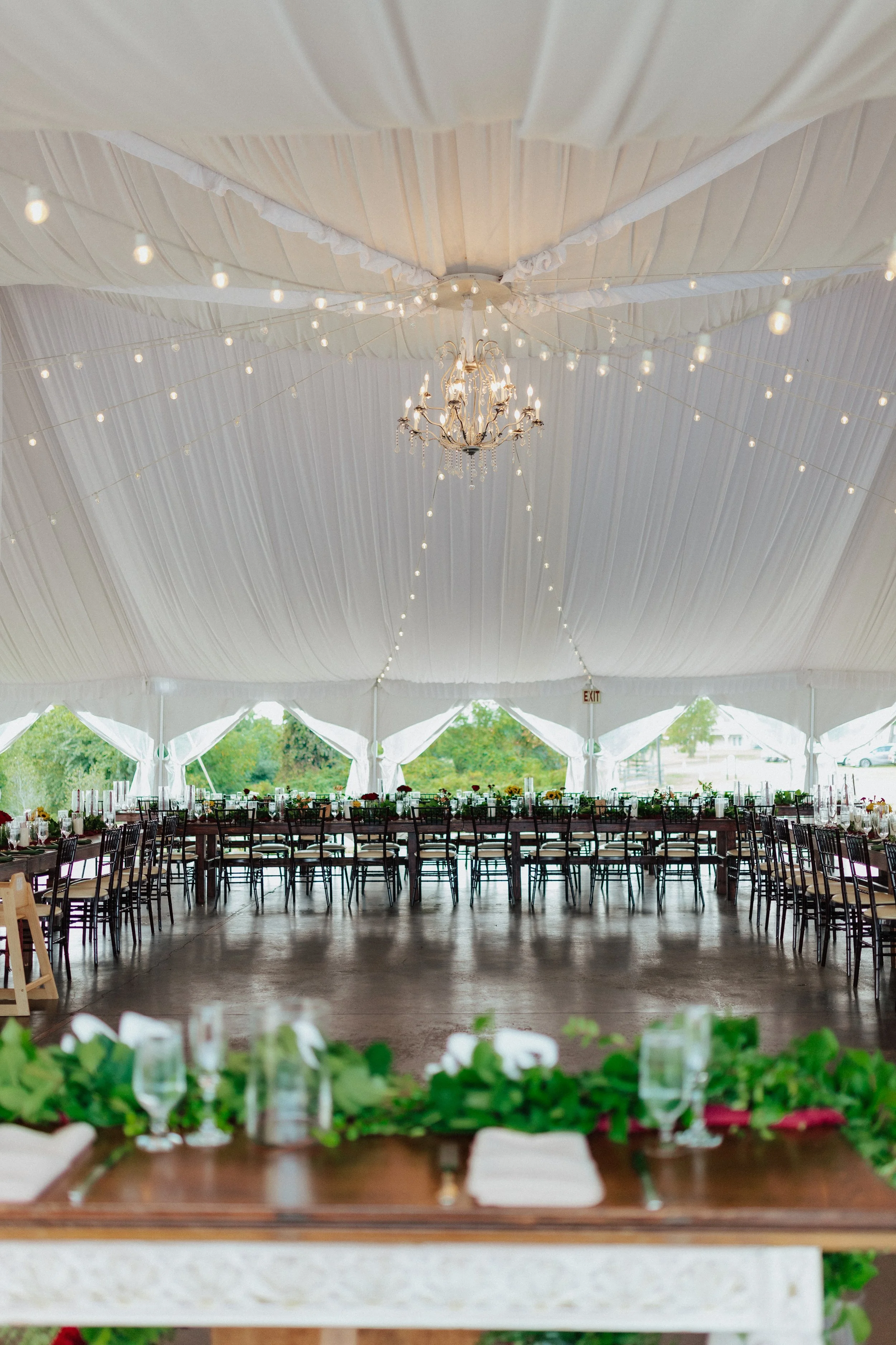 The interior of a large tent decorated for an event with a chandelier hanging from a white, draped ceiling and string lights. There are two rows of tables with chairs, set with glassware, candles, and floral centerpieces. The front of the image revea