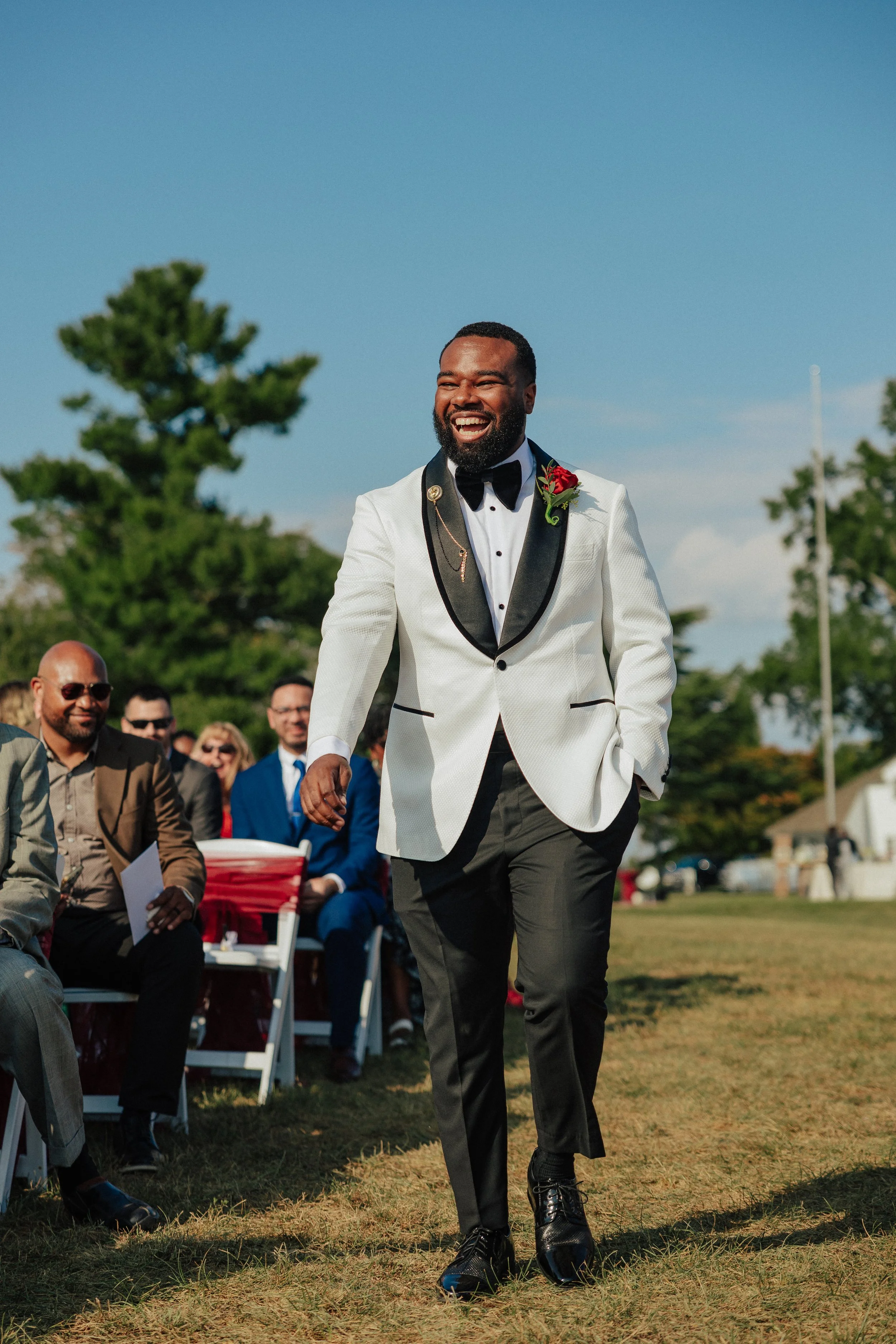 Man in a white tuxedo jacket with black trim and a red rose boutonniere, walking on grass at a formal outdoor event, smiling, with seated guests and green trees in the background.