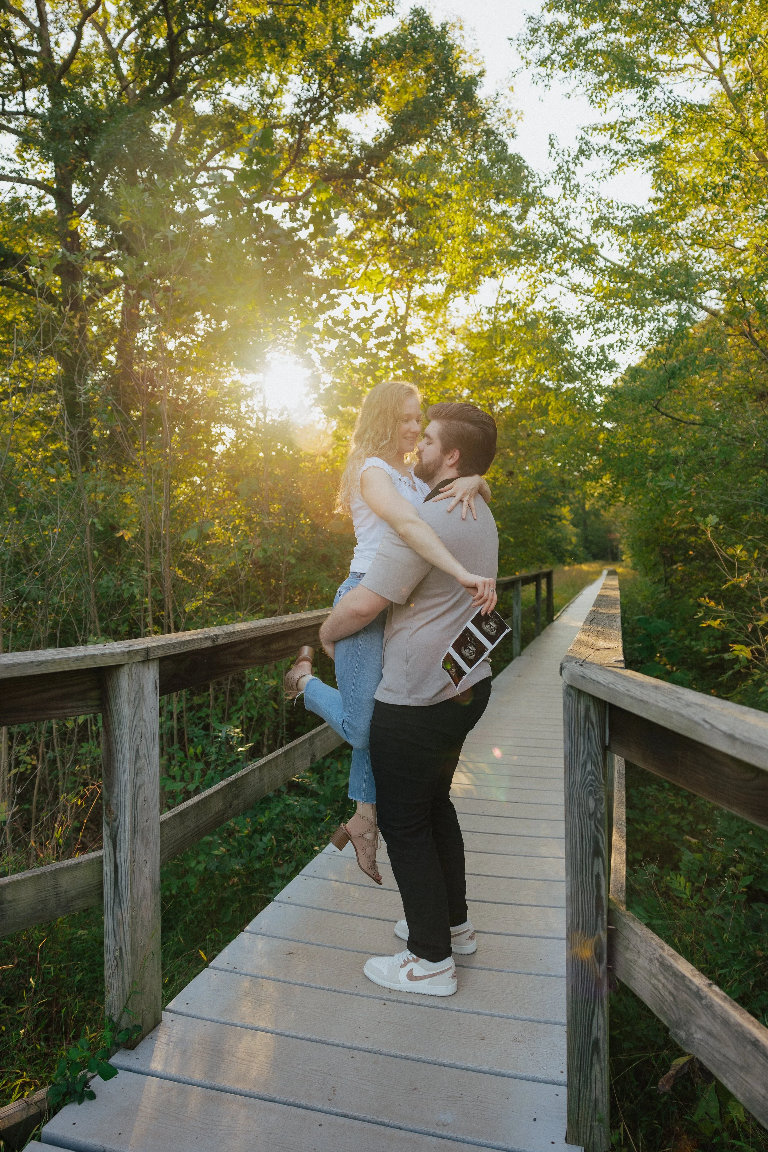 A young couple hugging on a wooden bridge in a lush green park during sunset, with trees and sunlight in the background.