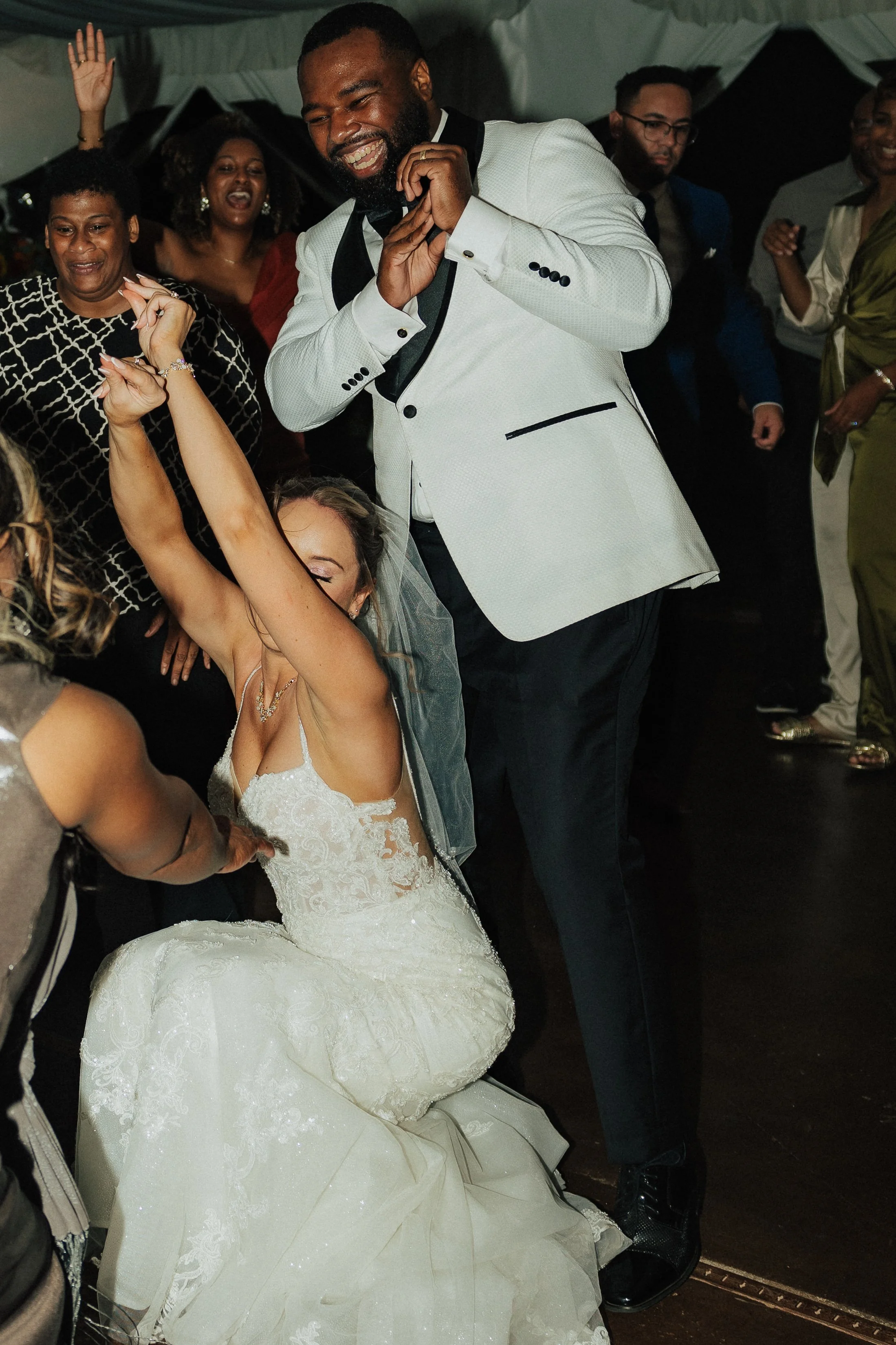 Celebration scene with a man in a white tuxedo dancing and smiling among a group of women, including a bride in a white wedding dress sitting on the floor, at a wedding or party.