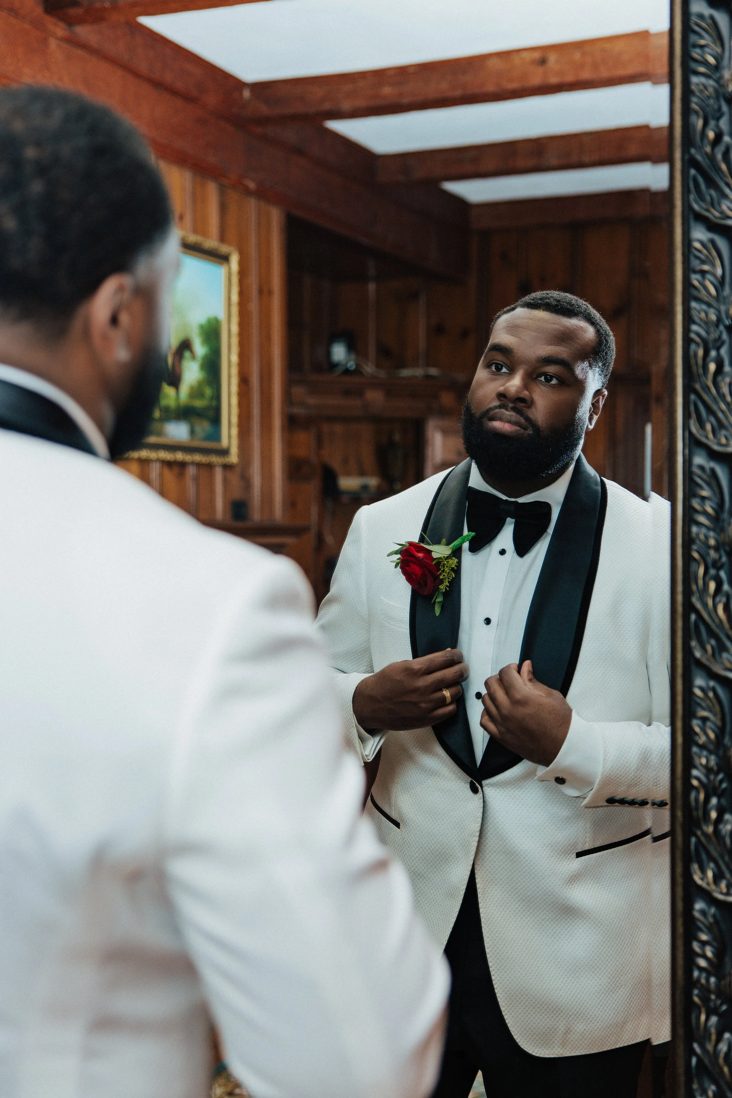 A man in a white tuxedo with a black lapel and a red rose boutonniere looking at his reflection in a mirror, adjusting his tuxedo, in a wood-paneled room.