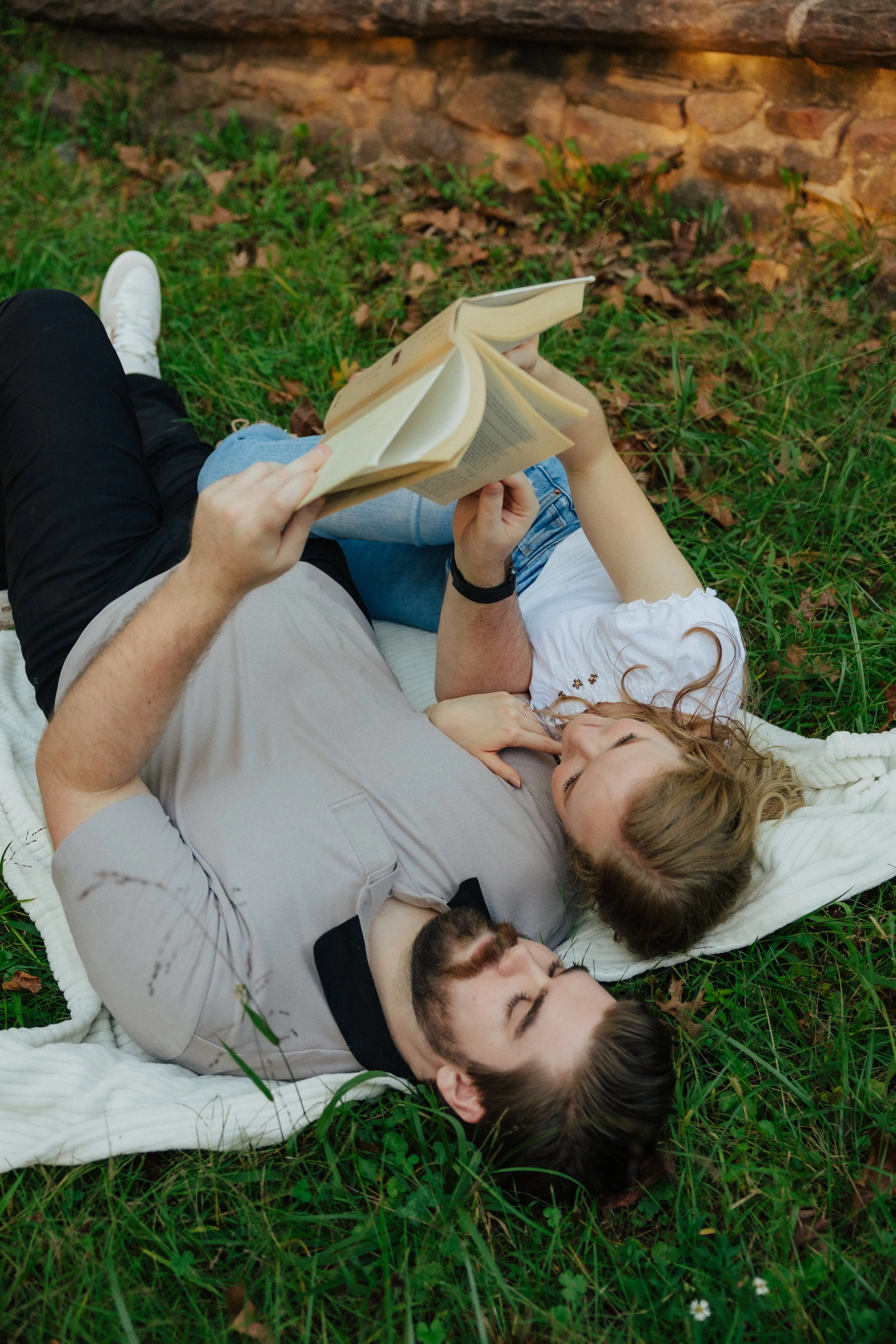 A group of four young adults lying on a blanket in a grassy area, reading books and relaxing outdoors.