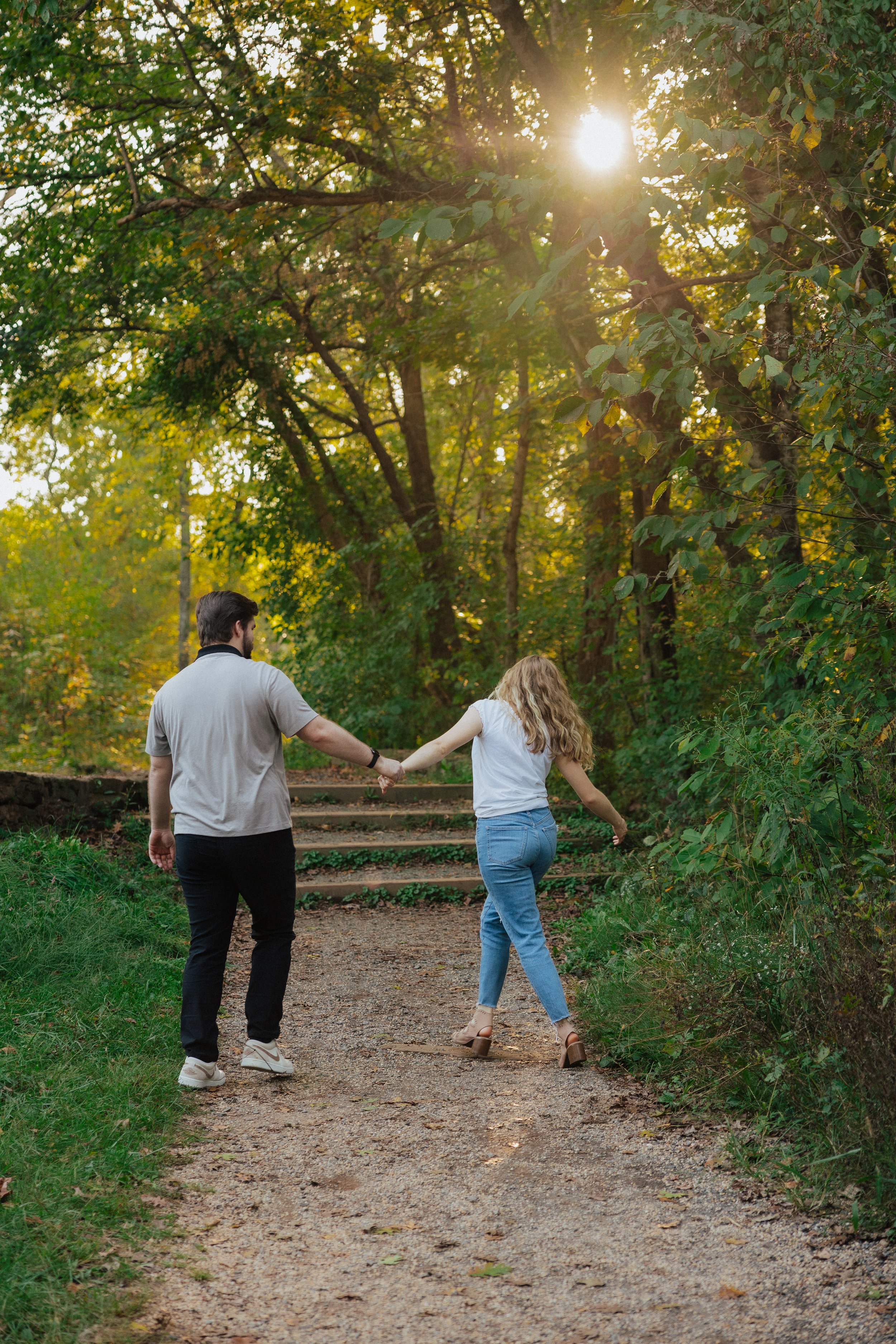 A couple walking hand in hand down a dirt trail in a woods with sunlight filtering through the trees.