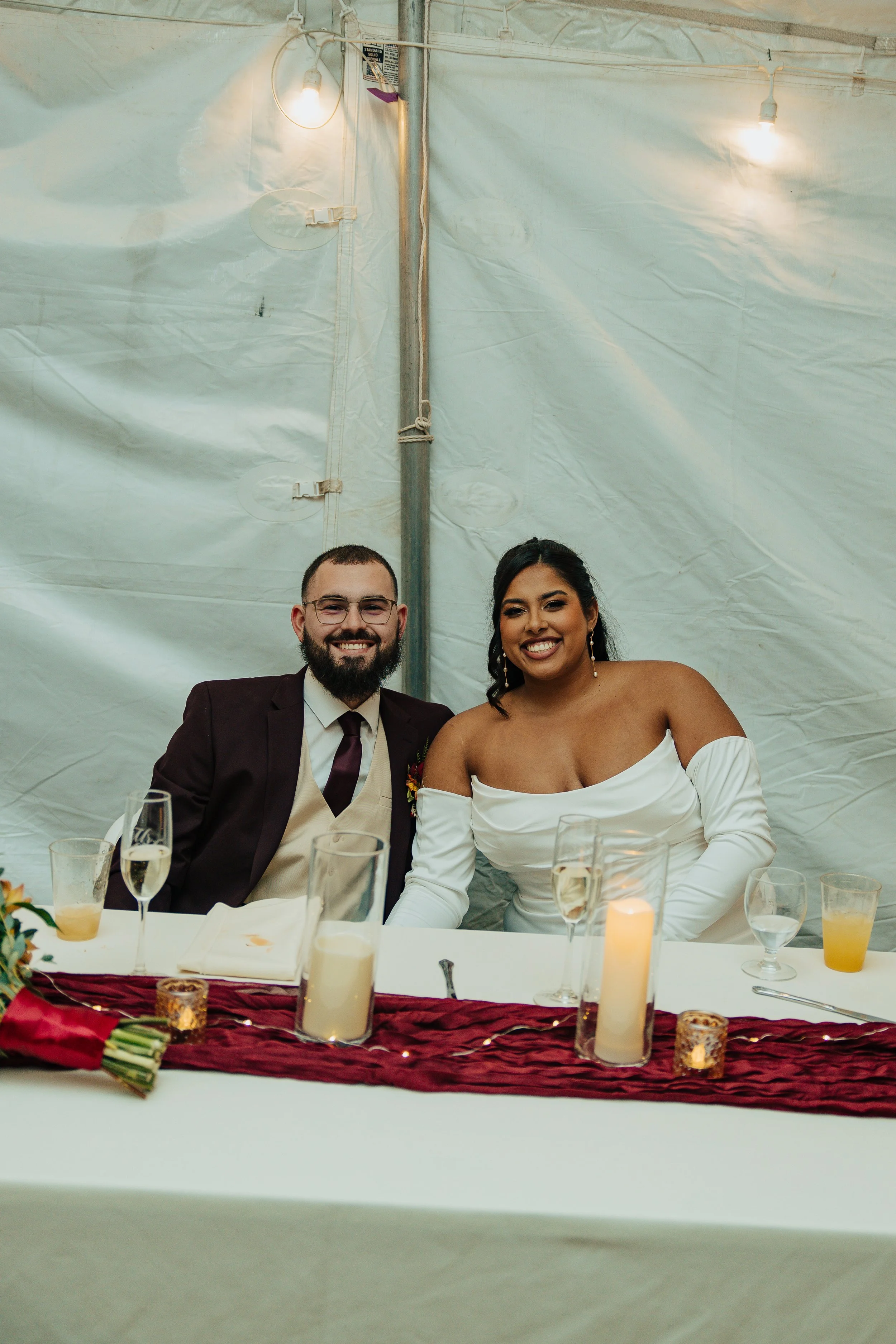 A man with glasses and a beard wearing a dark suit and a woman with dark hair in an off-the-shoulder white dress sitting at a wedding reception table decorated with candles, glasses of champagne, and floral arrangements. They are smiling in front of 