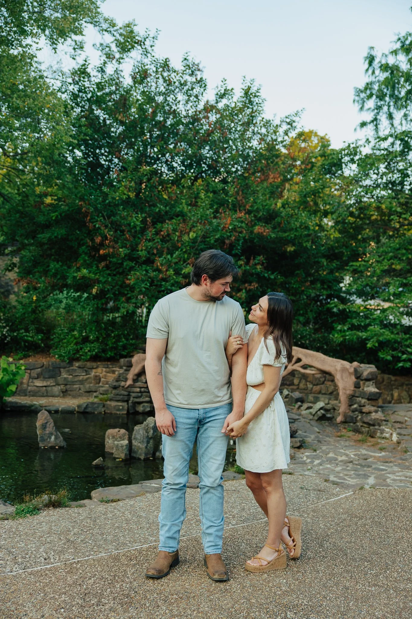 A couple standing near a pond surrounded by greenery, holding hands and looking into each other's eyes, with trees and stone features in the background.