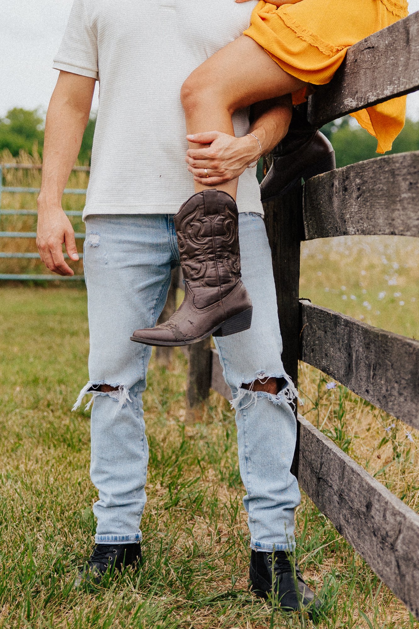 A person wearing a white shirt and ripped jeans is standing outdoors next to a wooden fence, while another person with a yellow dress and brown cowboy boots is sitting on the fence with their leg resting on it.
