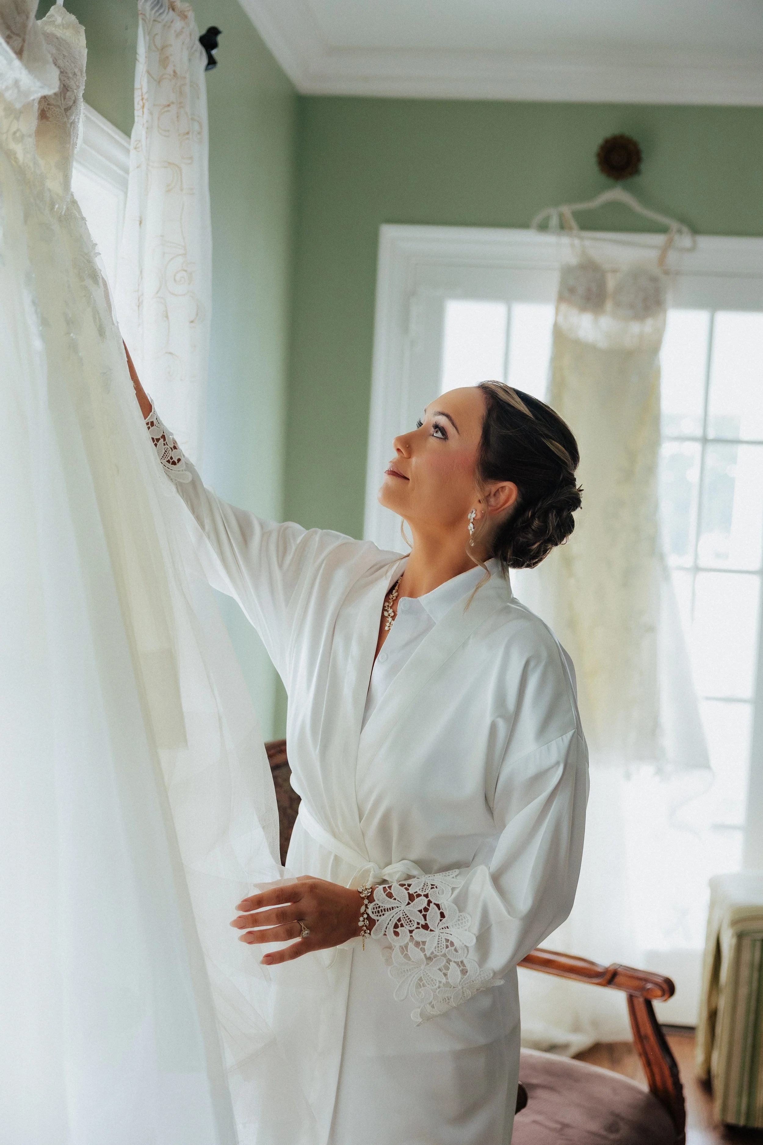 A woman in a white robe reaching for a wedding dress hanging near a window in a softly lit room.