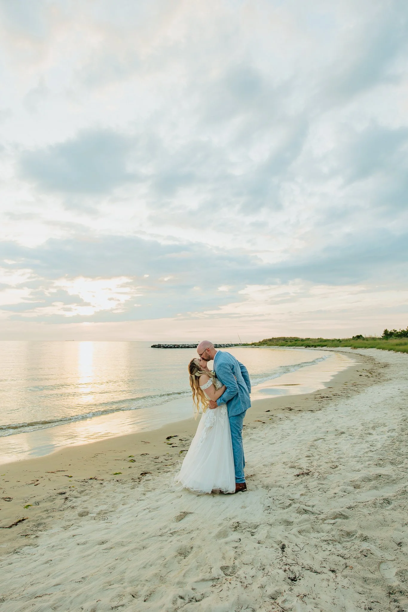 A couple sharing a kiss on the beach during sunset, with the woman in a white wedding dress and the man in a gray suit and glasses, near the shoreline with the sky filled with clouds.