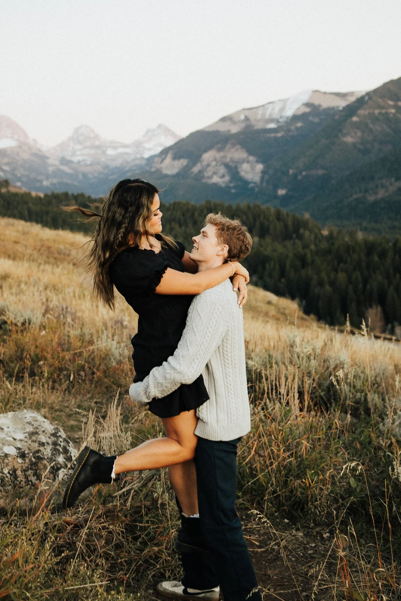 A young woman being lifted by a young man in a grassy, mountainous outdoor setting during daytime. The woman has long wavy hair and is wearing a black dress and black boots. The man has short hair and is wearing a light-colored sweater and black pants. There are mountains and a forest in the background.