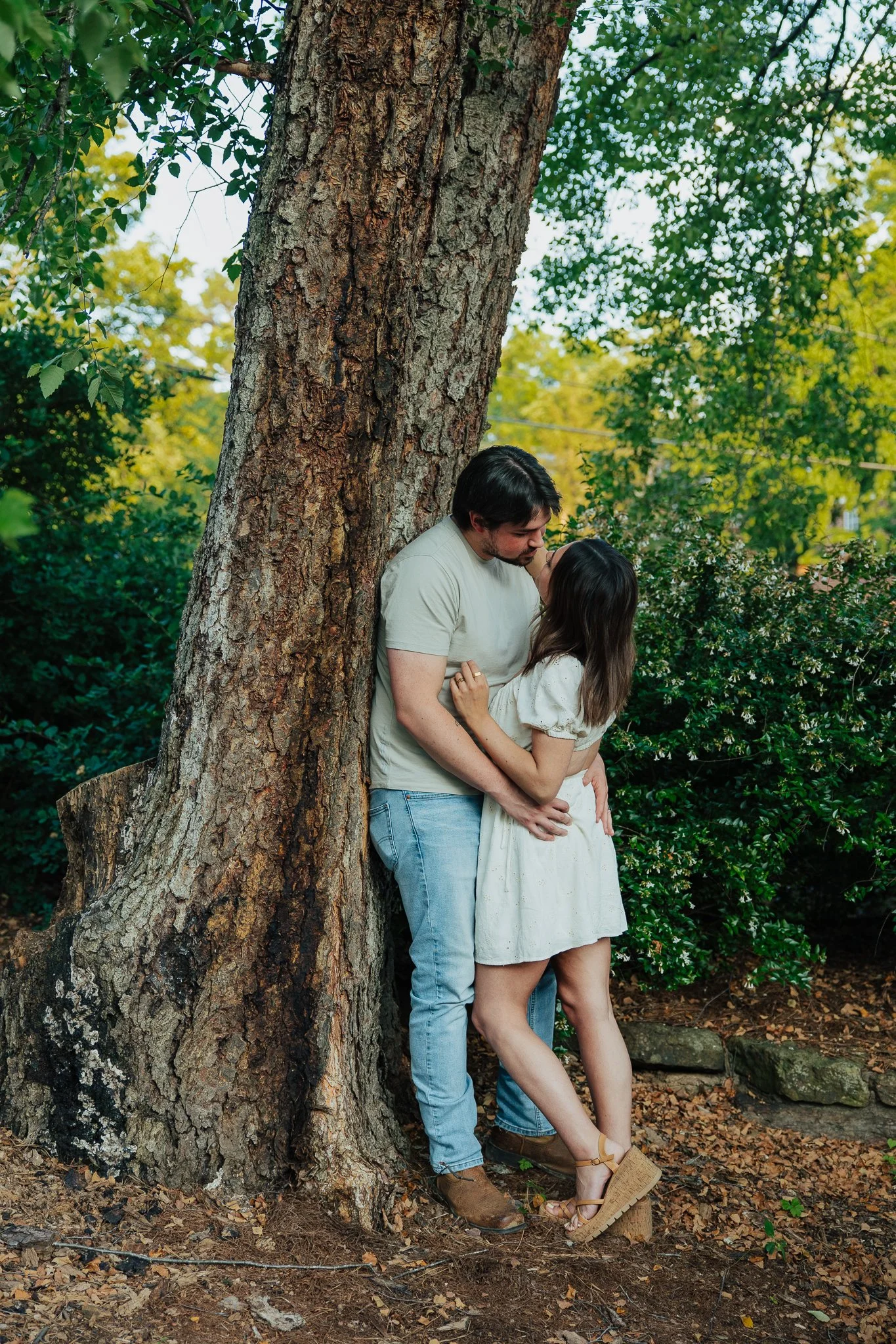 A couple embracing and sharing a kiss outdoors near a large tree surrounded by greenery and fallen leaves.