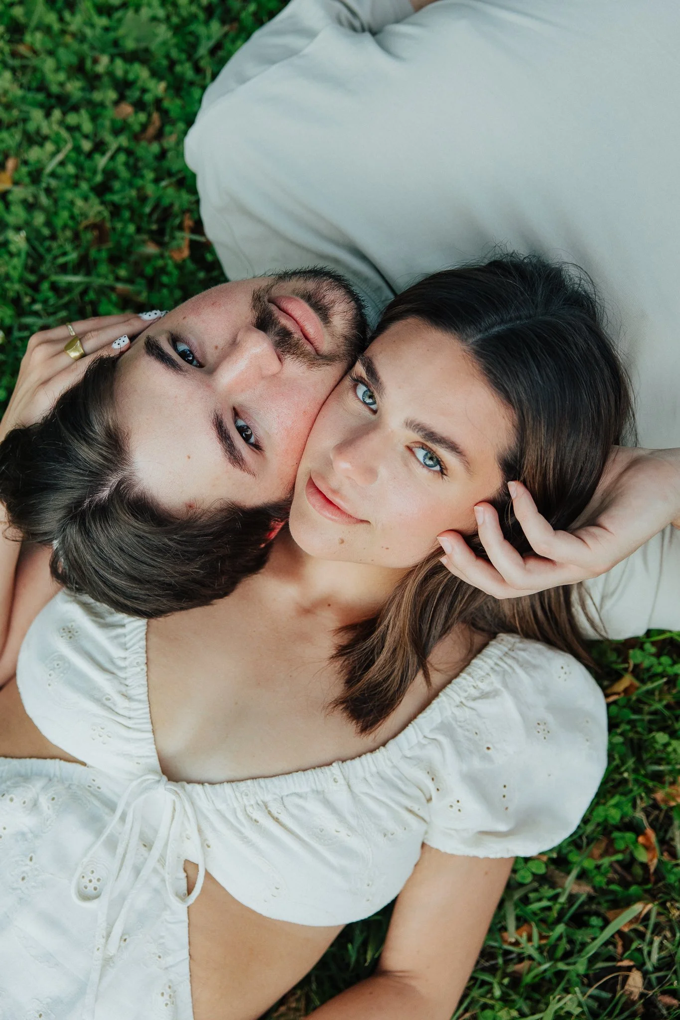 A woman and a man lying on the grass, very close or possibly hugging, with the woman’s head near the man’s face. The woman has long dark hair and blue eyes, and is looking at the camera. The man has dark hair, a beard, and is winking or closing one e