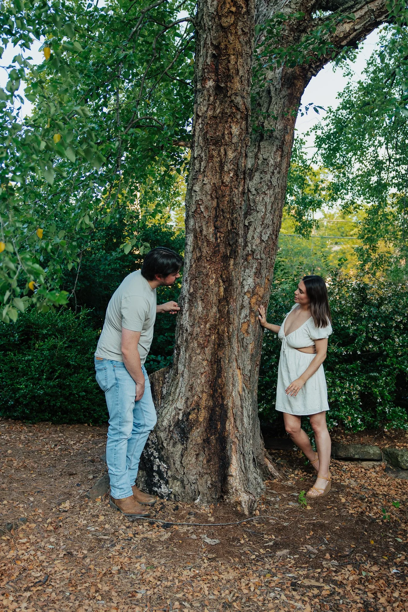 A man and woman are standing on opposite sides of a large tree trunk in a garden, looking at each other. The man is wearing a beige T-shirt, blue jeans, and brown boots, while the woman is in a white dress and beige high-heeled sandals. There are gre