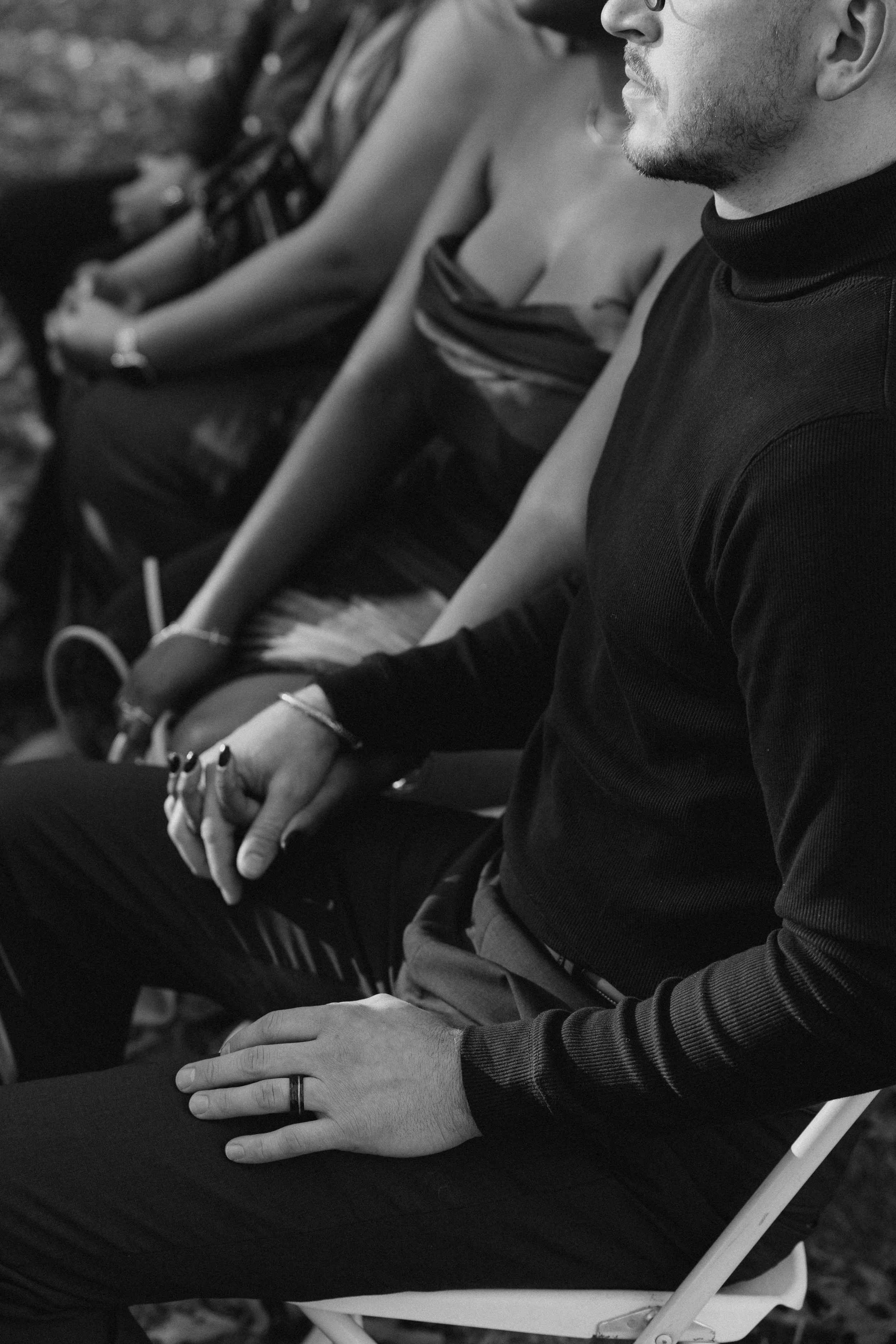 Black and white photo of two people sitting side by side, with focus on their hands and lower arms. One person wears a ring on their finger, while the other has a bracelet. Their faces are partially visible, showing one person with glasses and facial