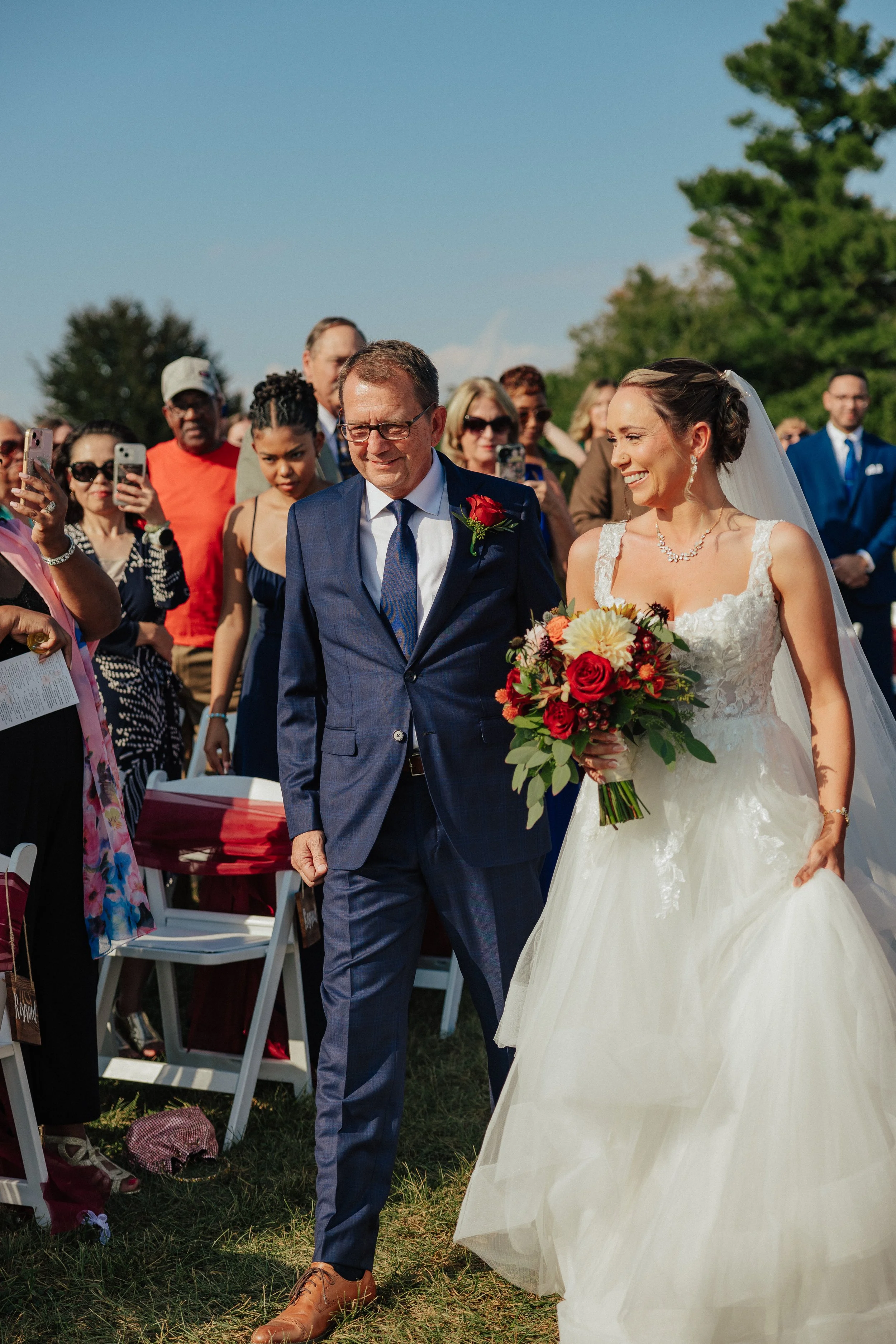 A bride in a white wedding dress holding a bouquet of flowers walking outside with a man in a blue suit, surrounded by wedding guests taking photos.