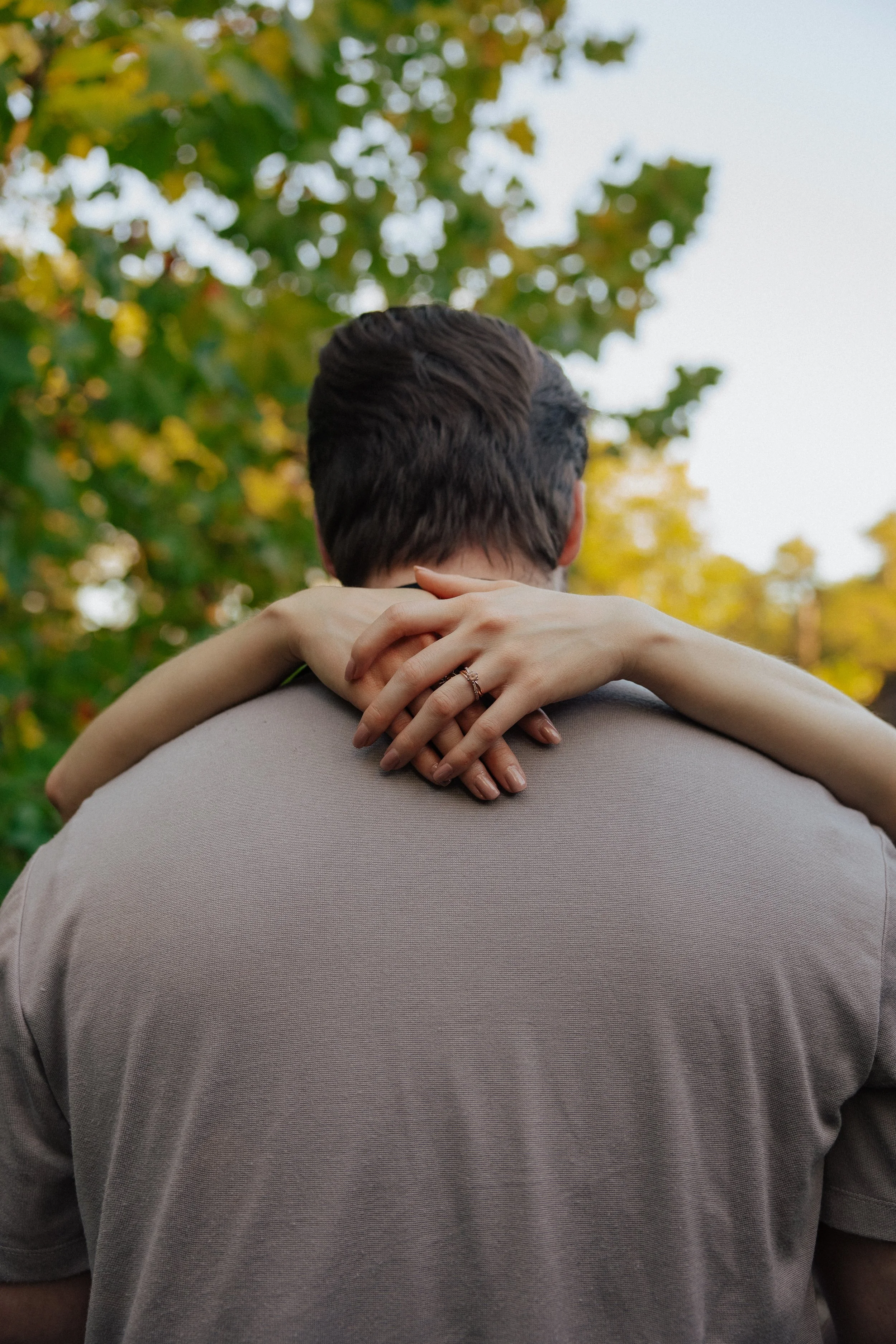 A person with short dark hair is sitting behind another person, wrapping their arms around their neck in an affectionate embrace outdoors, with blurred green and yellow autumn leaves in the background.
