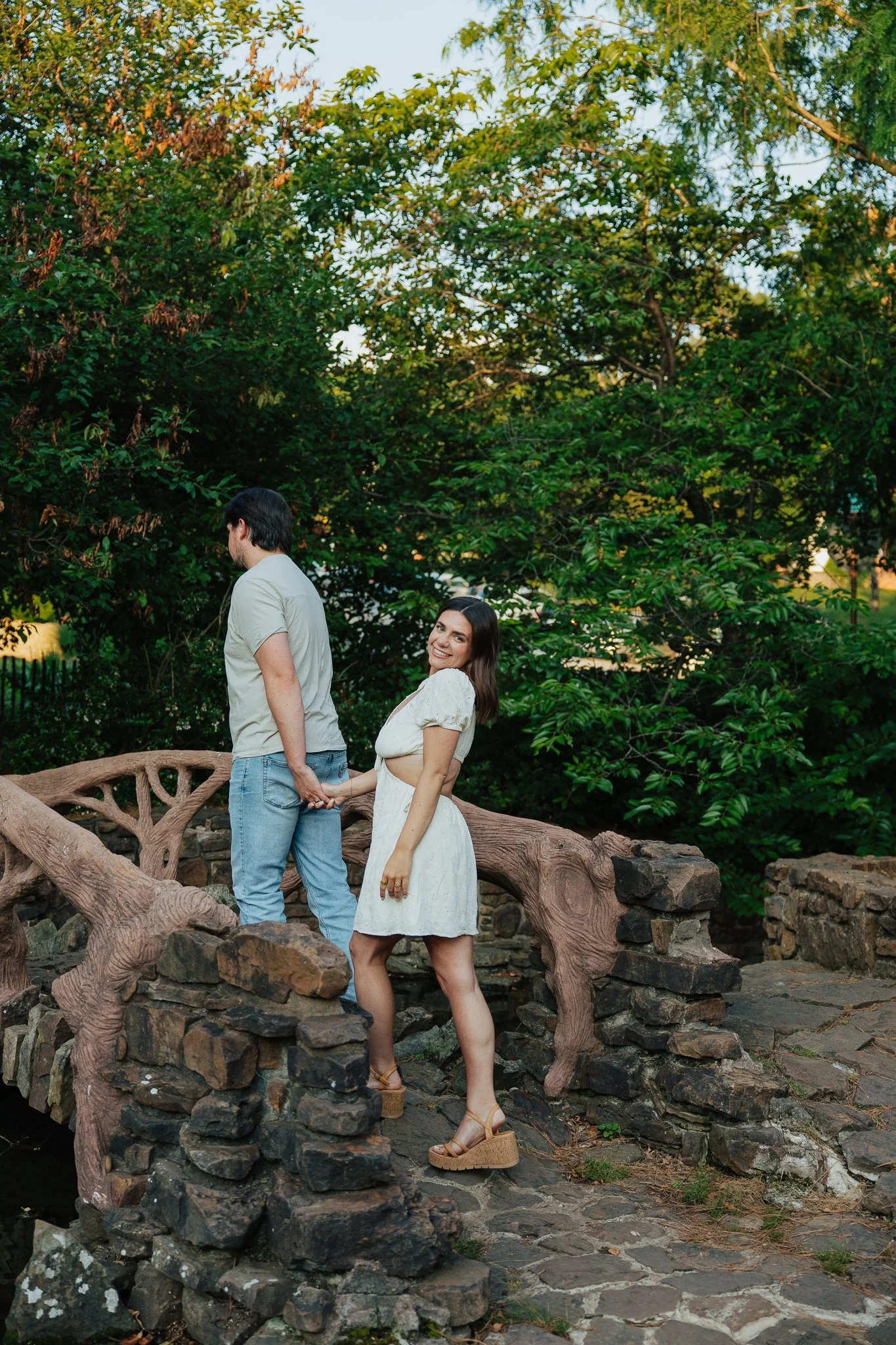 A woman in a white dress stands on a stone and wooden bridge outdoors, holding hands with a man in a light-colored shirt and jeans, with a lush green background of trees and foliage.