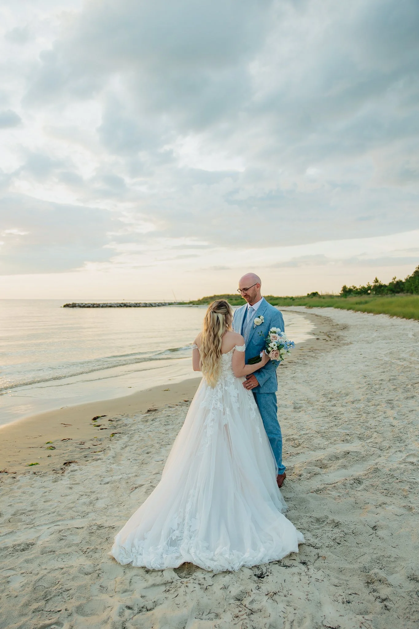 A bride and groom standing on a beach, holding hands and facing each other, with the bride in a white wedding gown and the groom in a light blue suit. The sky is cloudy and the ocean is in the background.
