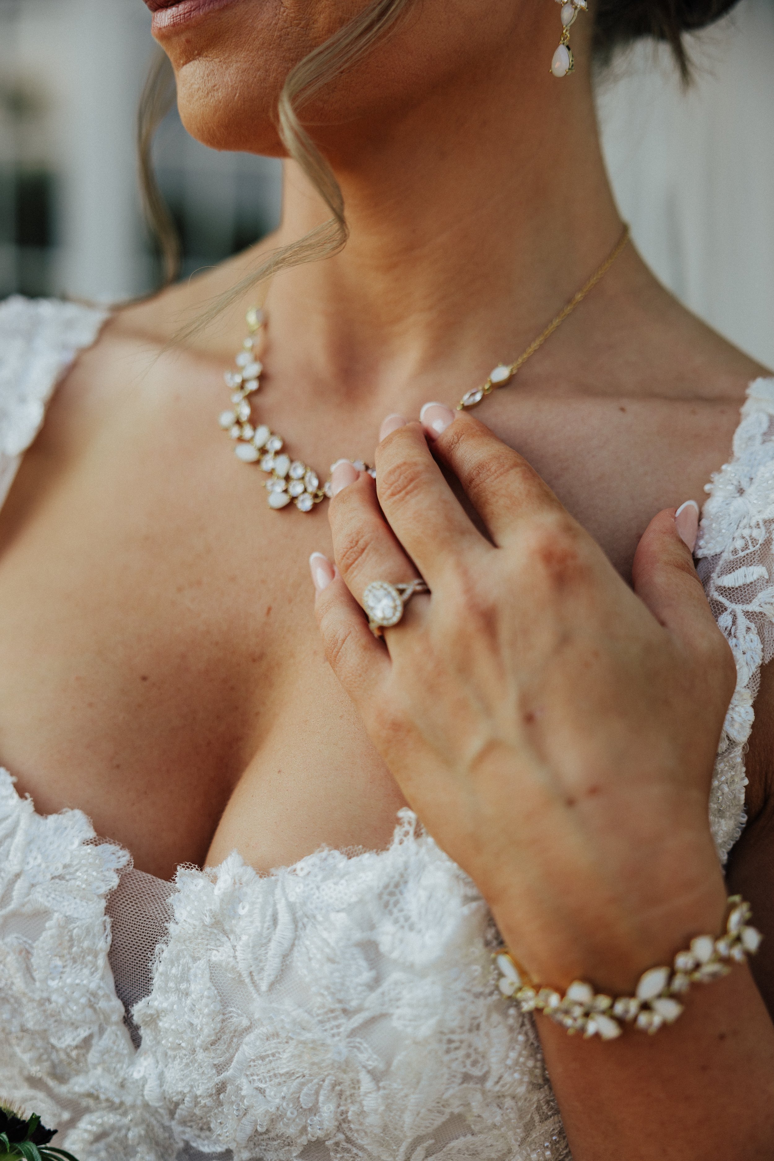 Close-up of a woman wearing a lace wedding dress and elegant jewelry, including a necklace, earrings, a ring, and bracelet, with her hand resting on her chest and a curl of hair near her face.