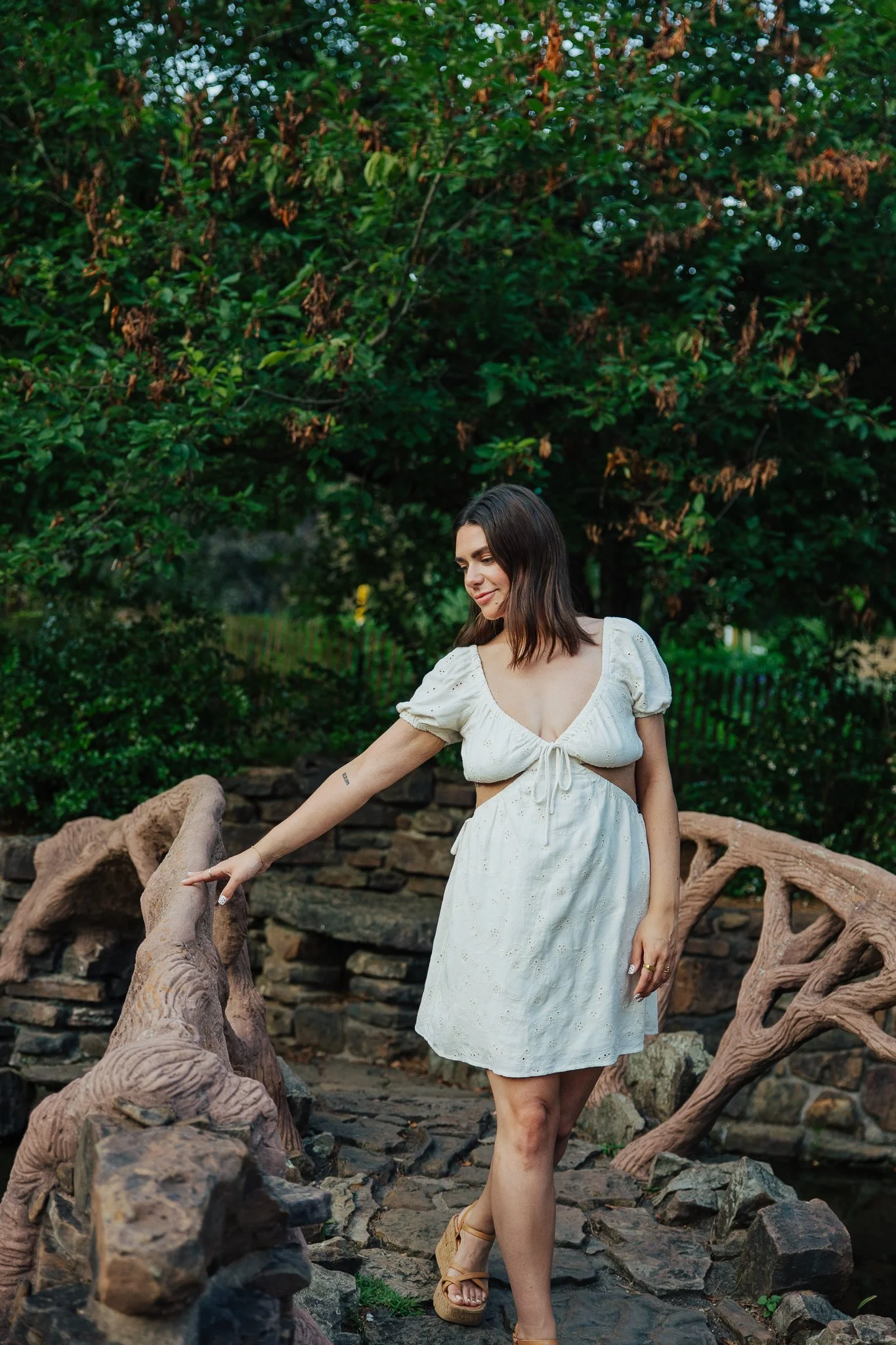 A woman in a white dress with puffed sleeves and a lace-up front walking on a rocky path under a large leafy tree, holding a unique wooden railing with natural curves.