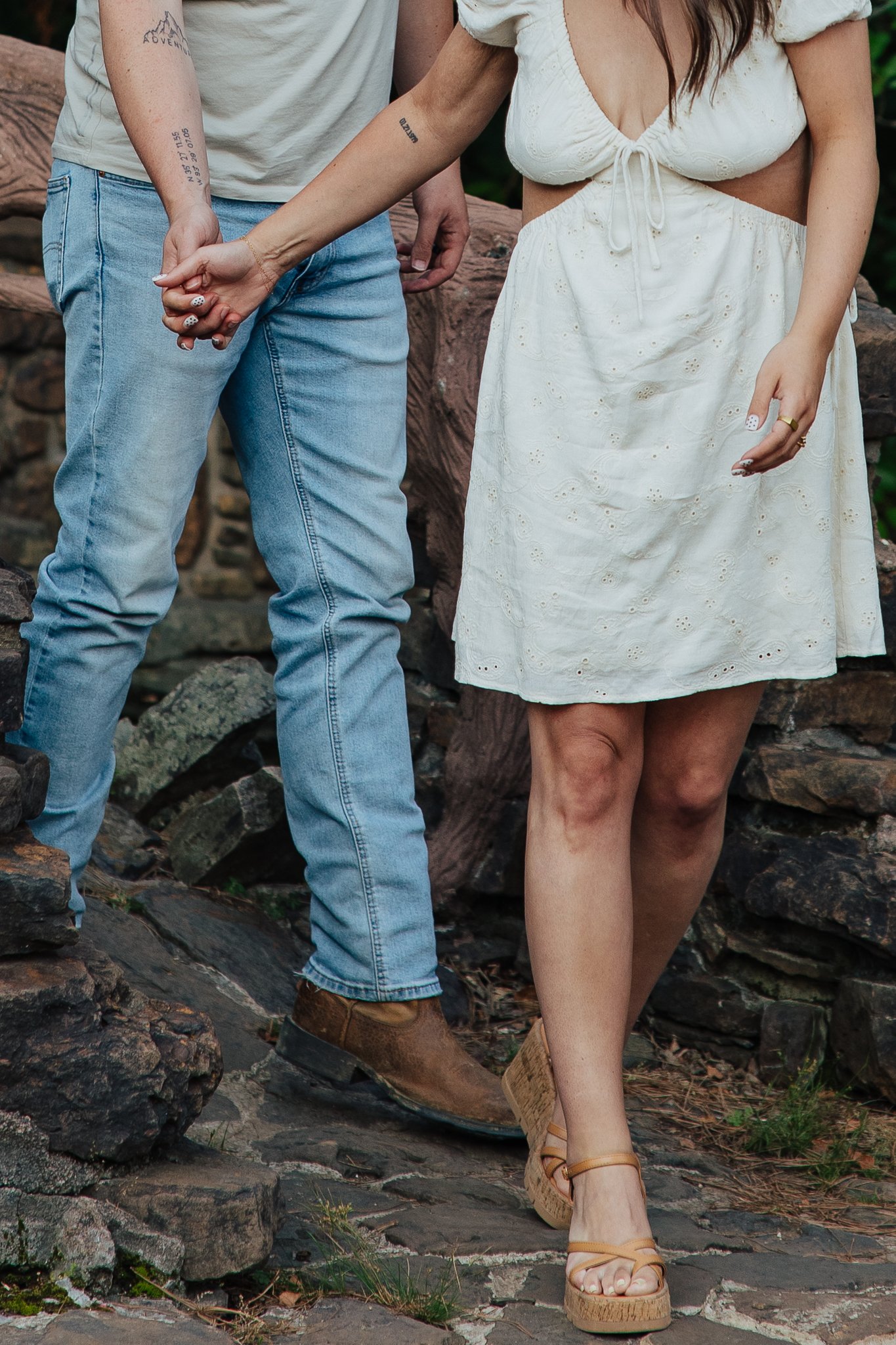 A man and woman holding hands, standing on a rocky outdoor surface. The woman is wearing a white lace dress and platform sandals, and the man is dressed in jeans and brown boots.