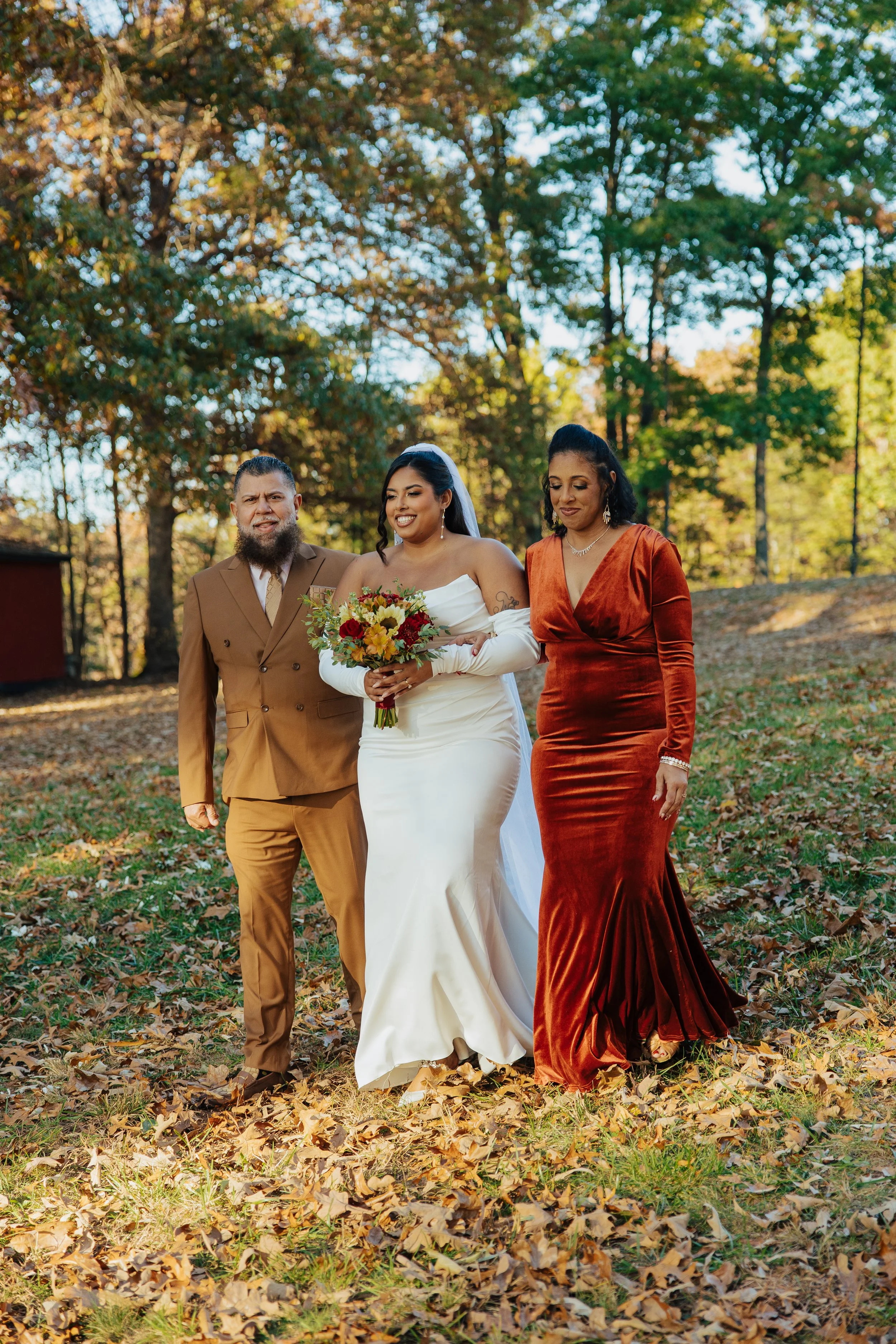 A bride and her parents walking outdoors in a park with fall foliage, the bride in a white gown holding a bouquet, and her parents in formal attire, the man in a brown suit and the woman in a rust-colored velvet dress.
