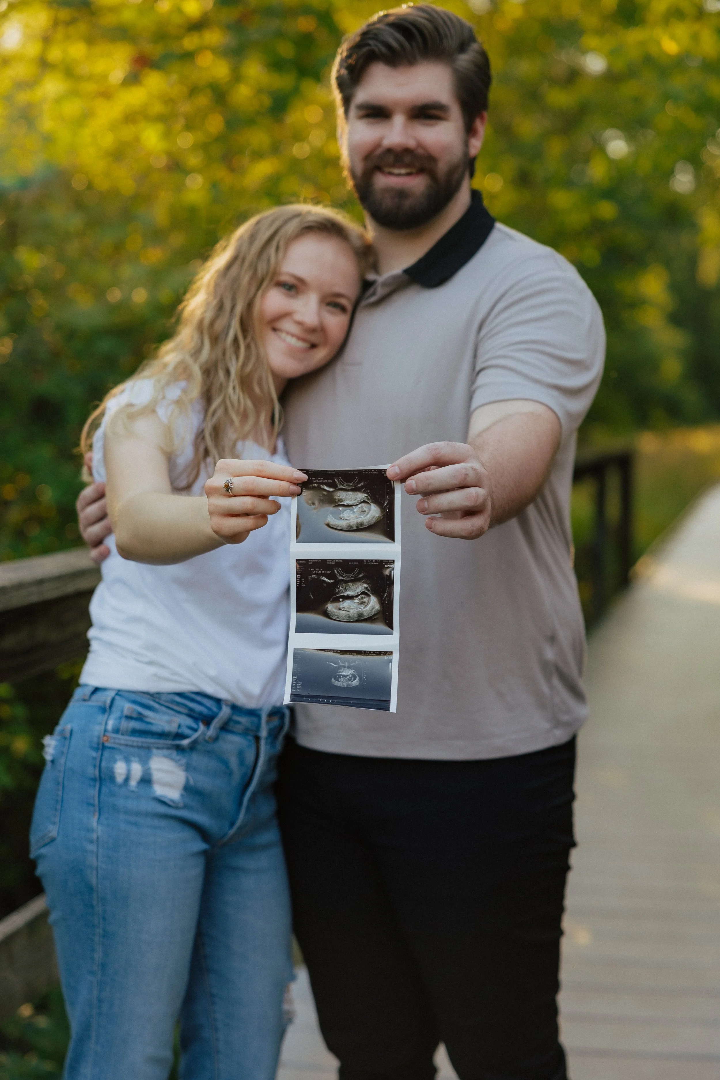A smiling woman and man standing outdoors, holding ultrasound pictures indicating pregnancy.