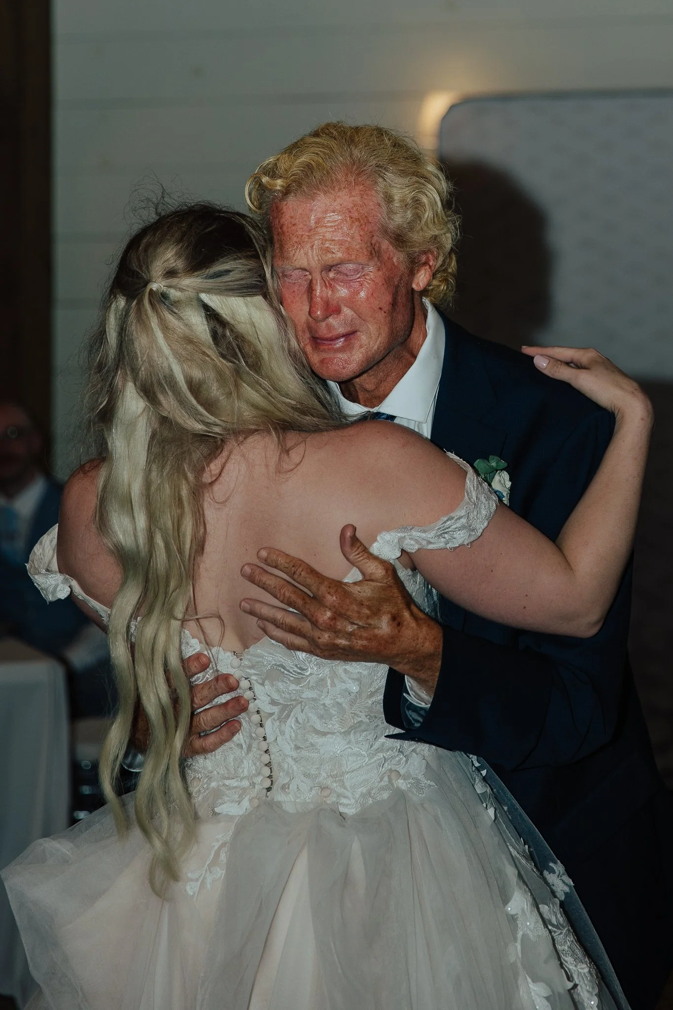 A bride and groom sharing a wedding dance, with the bride dressed in a white lace wedding gown and the groom wearing a dark suit, embracing each other closely during their wedding reception.