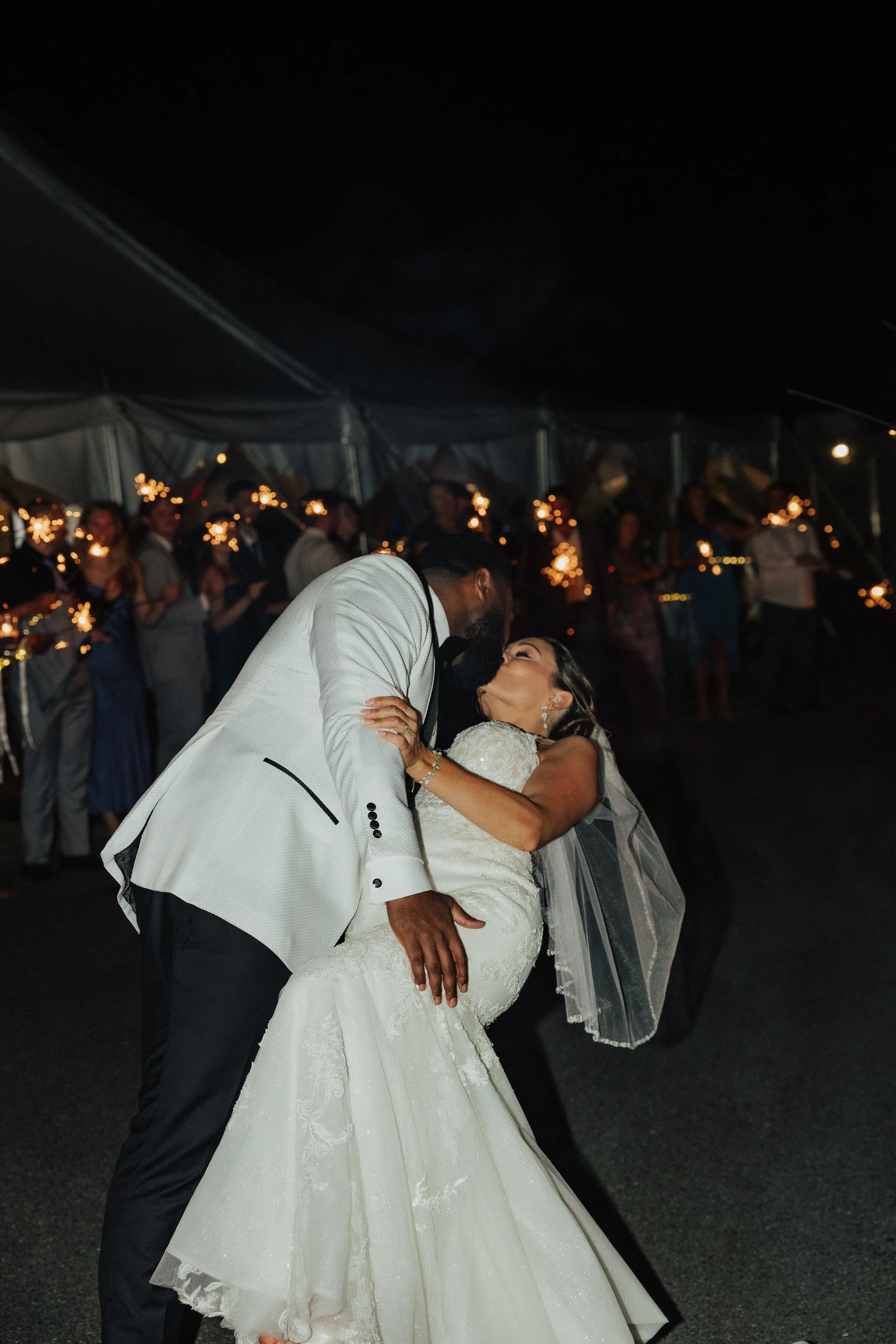 A bride and groom share a dance at their wedding reception, with guests holding sparklers in the background under a large tent at night.