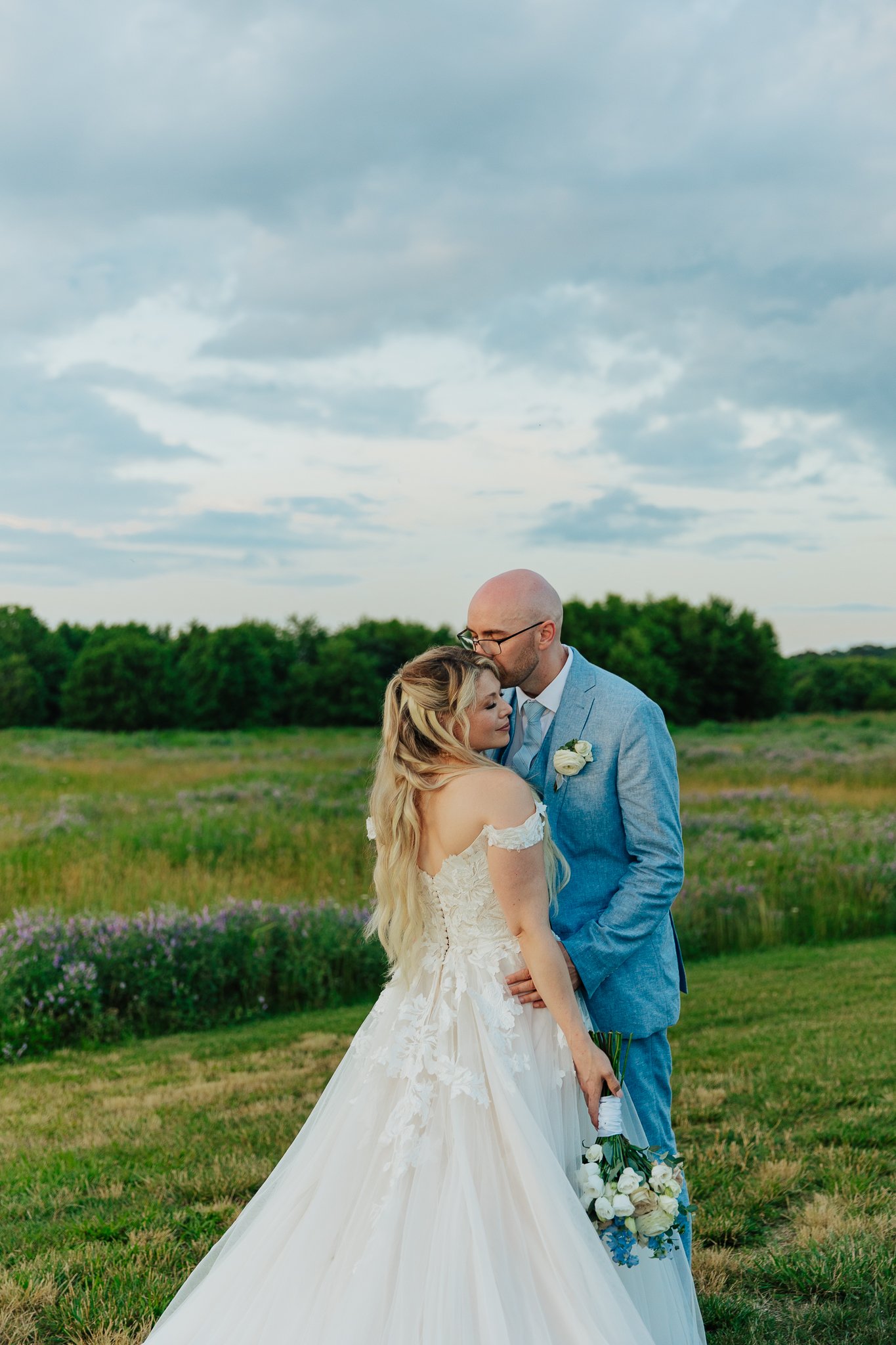 A bride and groom standing close together outdoors in a field, sharing a tender moment, with the groom gently kissing the bride on the forehead. The bride is holding a bouquet of white and blue flowers, and both are dressed in wedding attire, with the bride in a white wedding gown and the groom in a light blue suit. Overcast sky with clouds and greenery in the background.