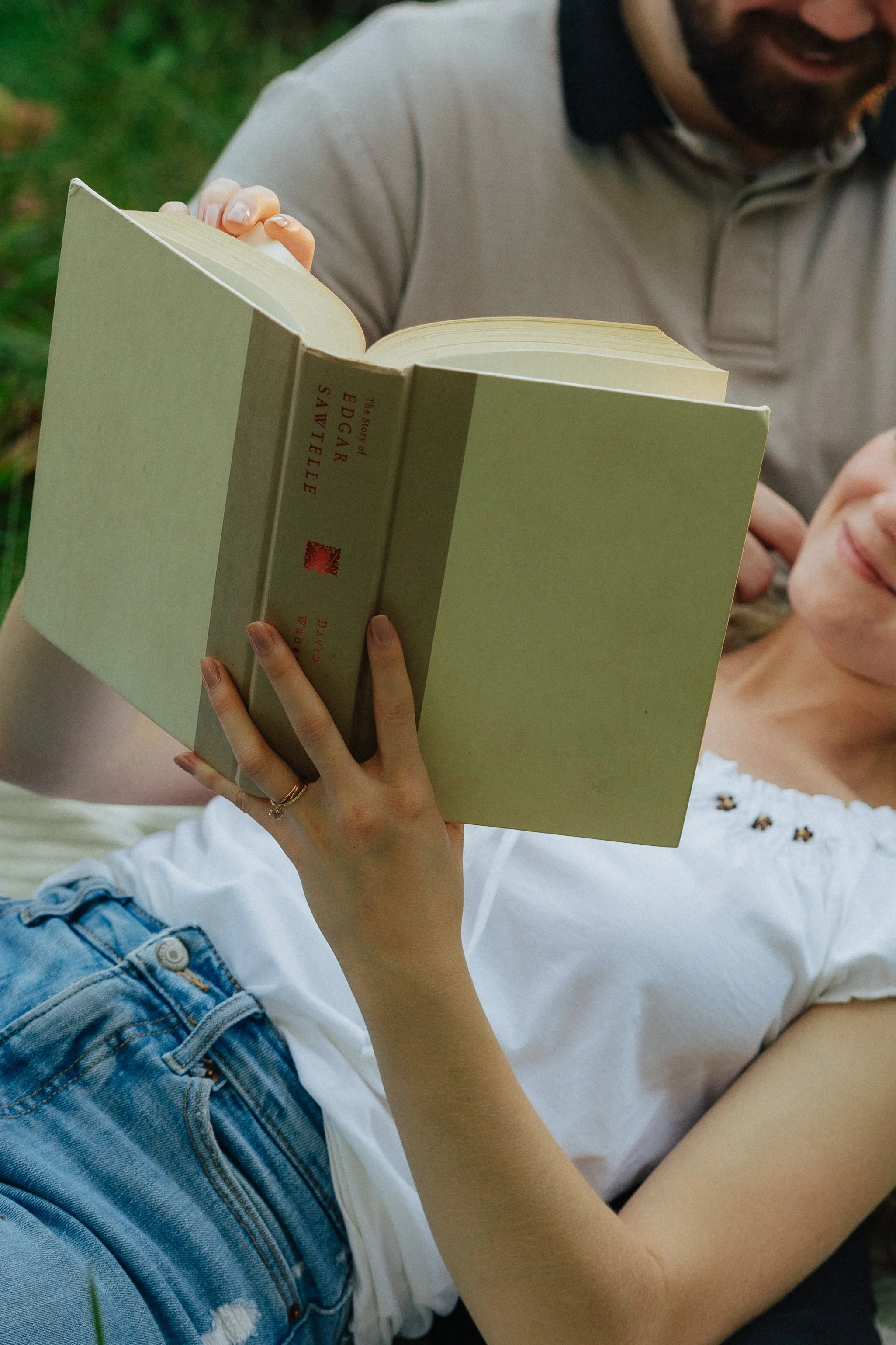 A person holding a book titled "The Story of Edgar Sawtelle" while sitting outdoors. The person is wearing a white shirt with embroidery and denim shorts.