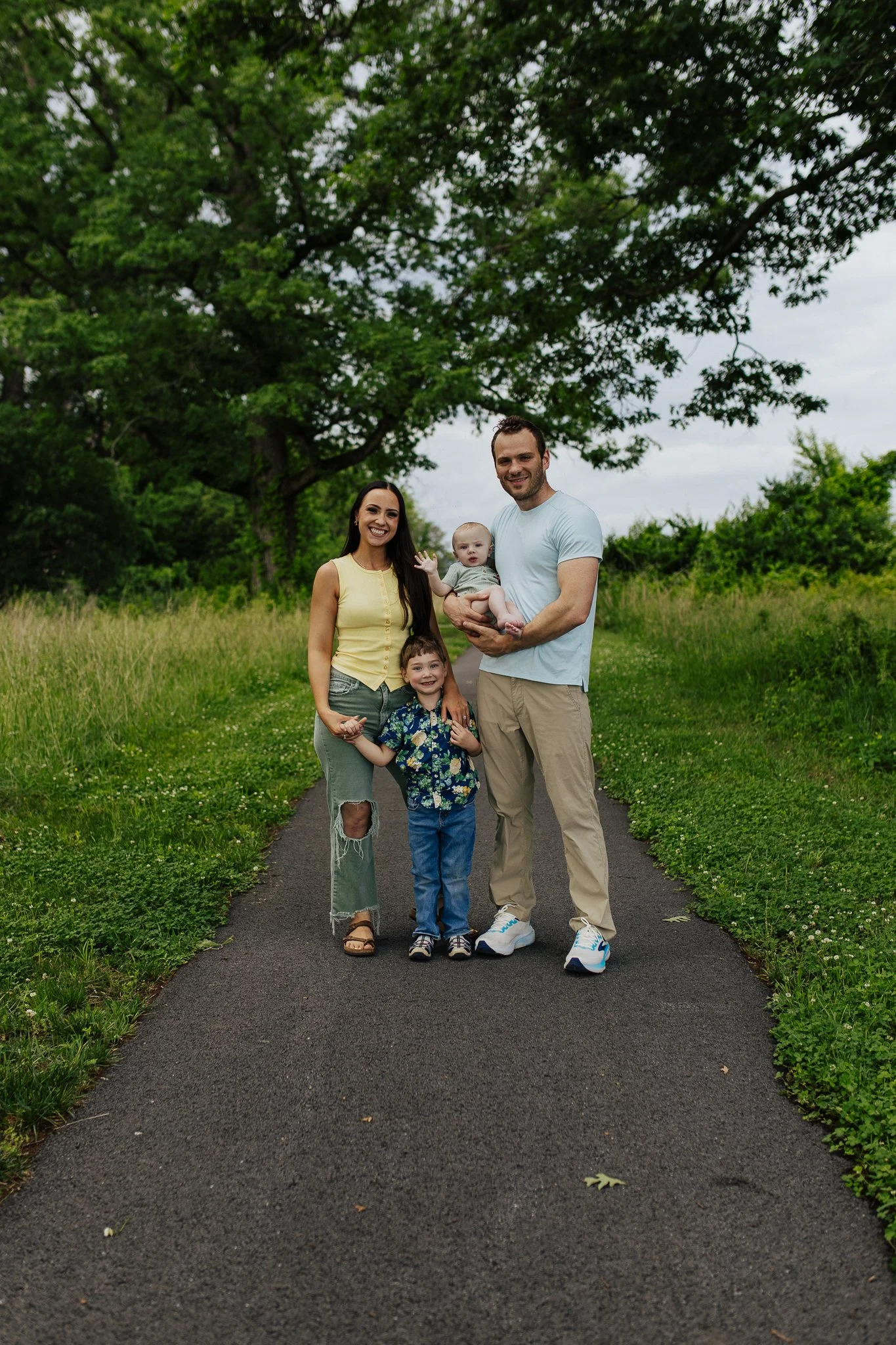 A family of five standing on a paved path in a park with green grass and trees. The family includes a woman, a man holding a baby, a young boy, and a young girl. They are smiling and enjoying a day outdoors.