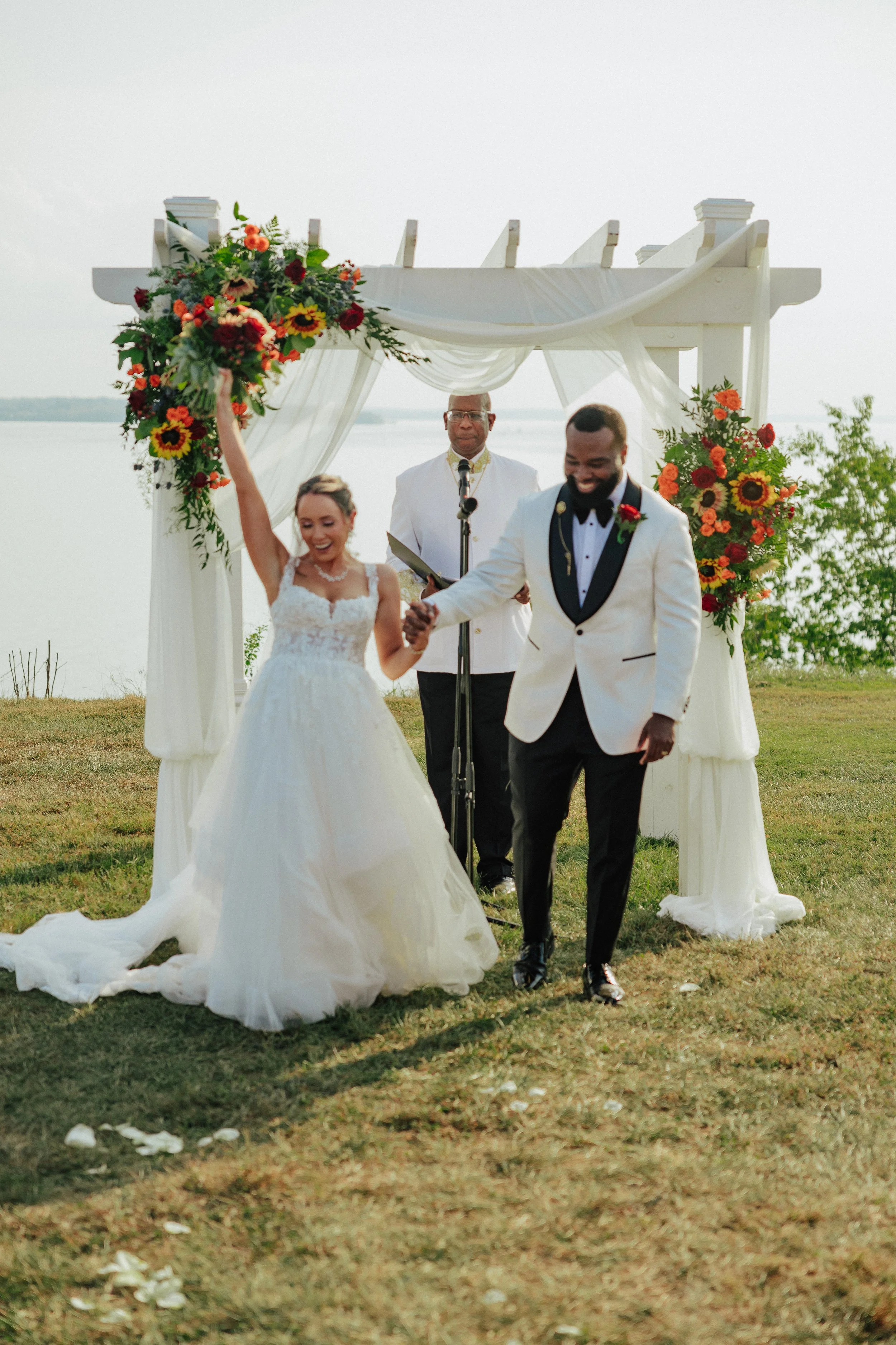 A bride and groom holding hands and smiling during their outdoor wedding ceremony near a lake. The bride is wearing a white wedding dress and the groom is in a white tuxedo with black lapels. An officiant stands behind them, and the ceremony is under