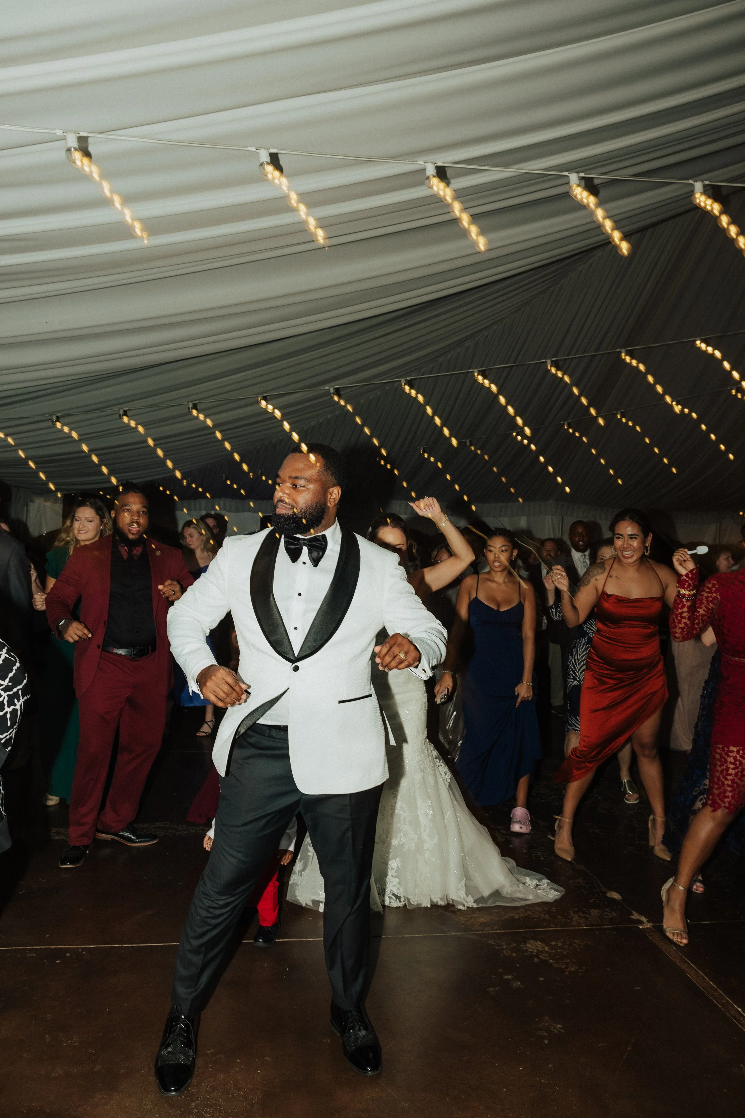 People dancing at a celebration or wedding reception inside a decorated tent with string lights. A man in the foreground is dressed in a tuxedo with a white jacket and black lapels, dancing with others around him, including a bride in a white gown.