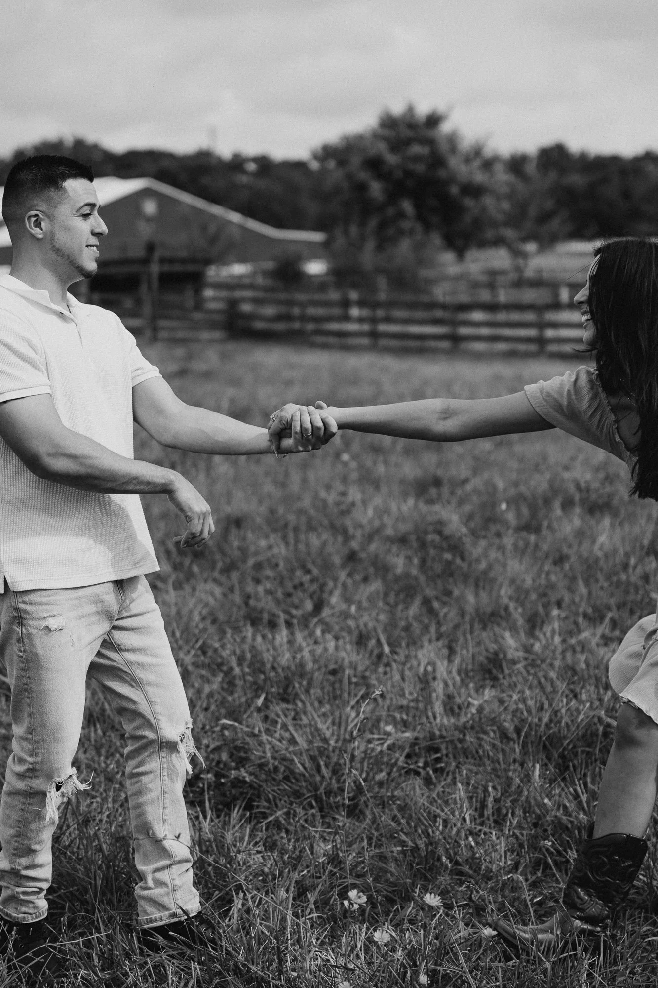 A young man and woman are outside in a grassy field, holding hands and smiling at each other. The man is standing while the woman is crouching slightly. They are shaking hands or holding hands with extended arms. A wooden fence and trees are in the b