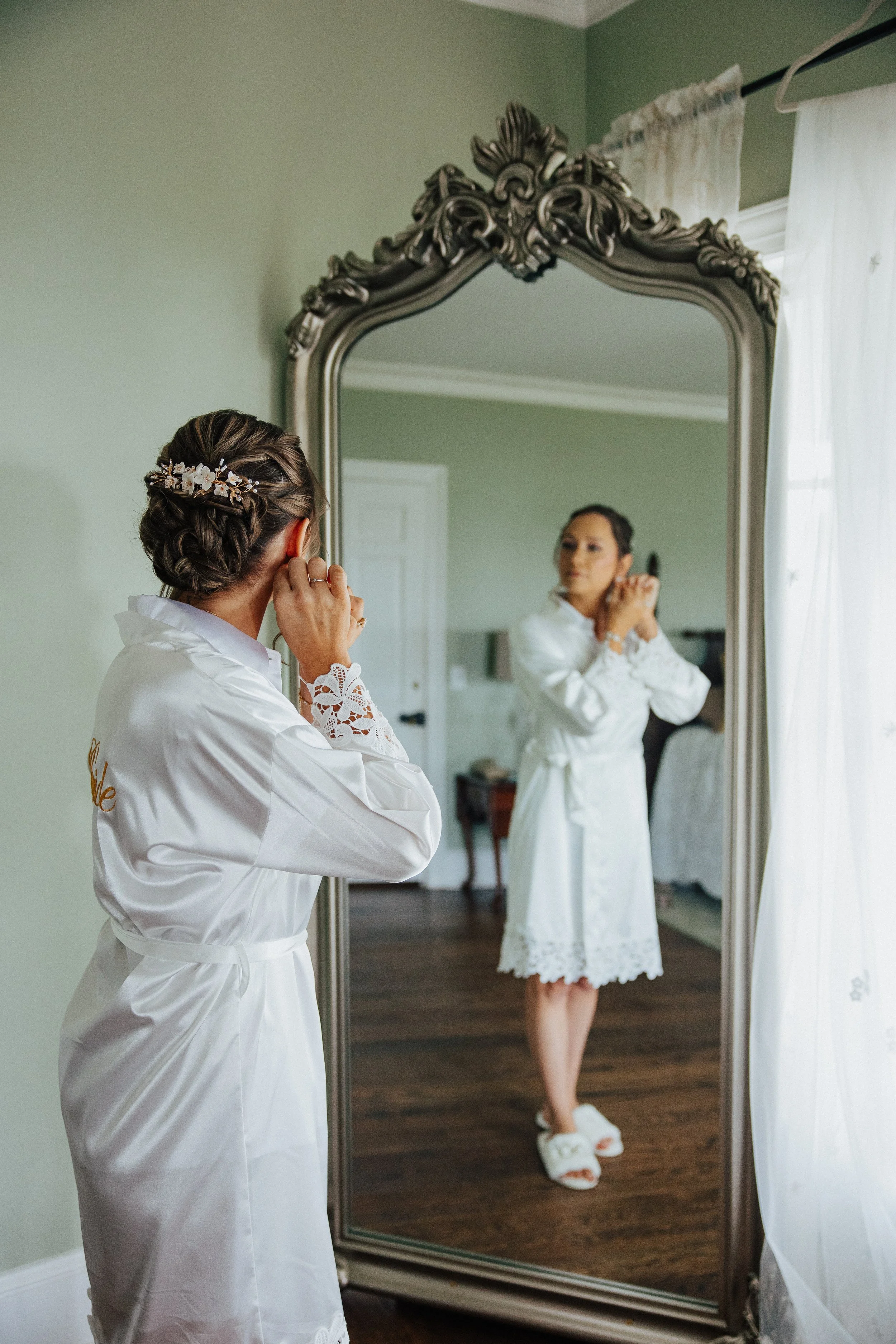 A woman with styled hair and a floral hair accessory is looking at herself in a large ornate mirror while adjusting her earrings. She is wearing a white robe with lace details and is in a room with a wooden floor and white curtains.