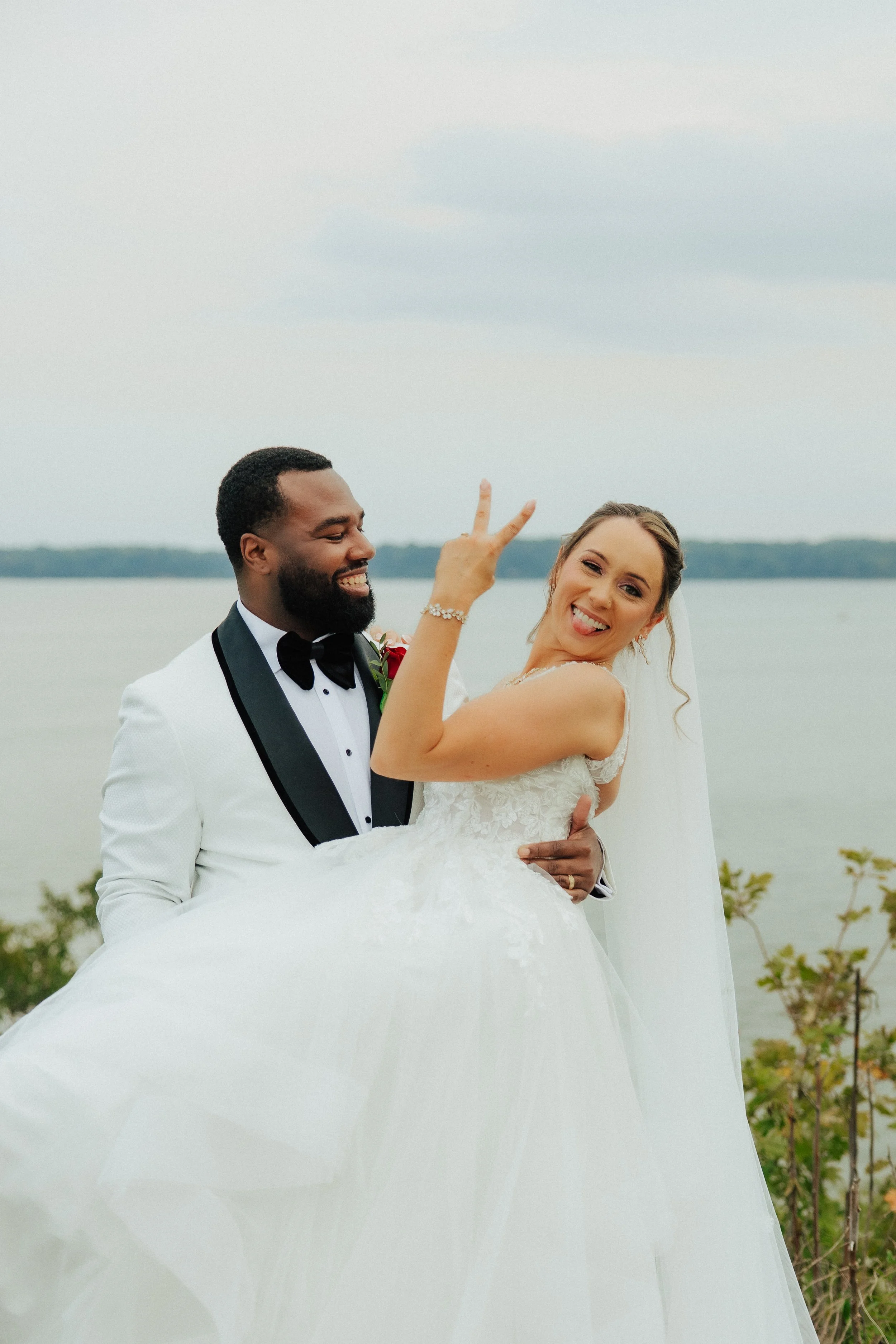 A happy interracial couple dressed in wedding attire, the groom in a white tuxedo with black lapel and bow tie, and the bride in a white lace wedding gown with a veil, are outdoors near water, with the groom holding the bride in his arms as she playf
