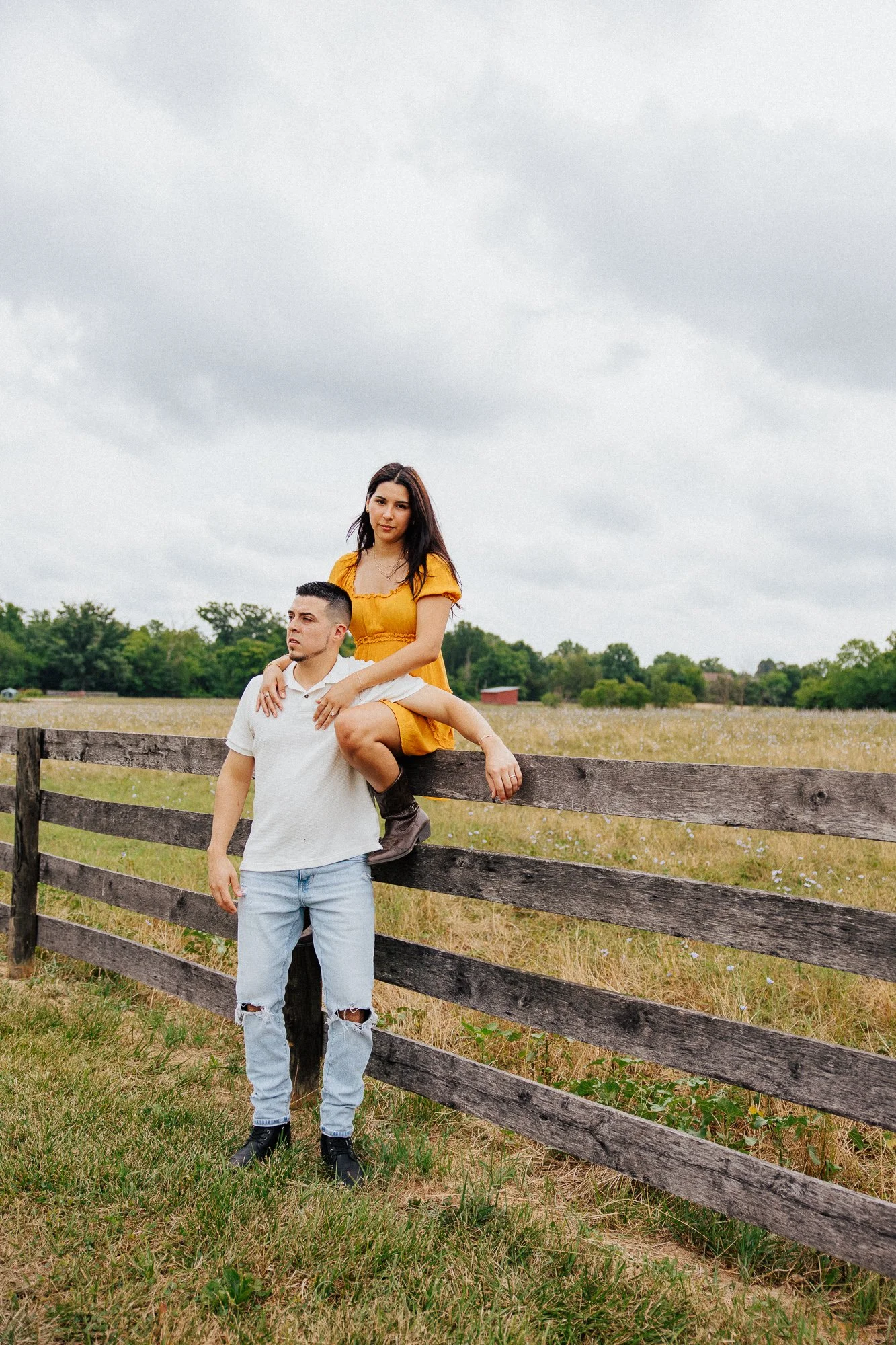 A man and a woman posing outdoors near a wooden fence in a field with trees and a cloudy sky in the background. The woman is sitting on the fence with one leg over and the man is standing beside her, holding her leg. The woman is wearing a yellow dre