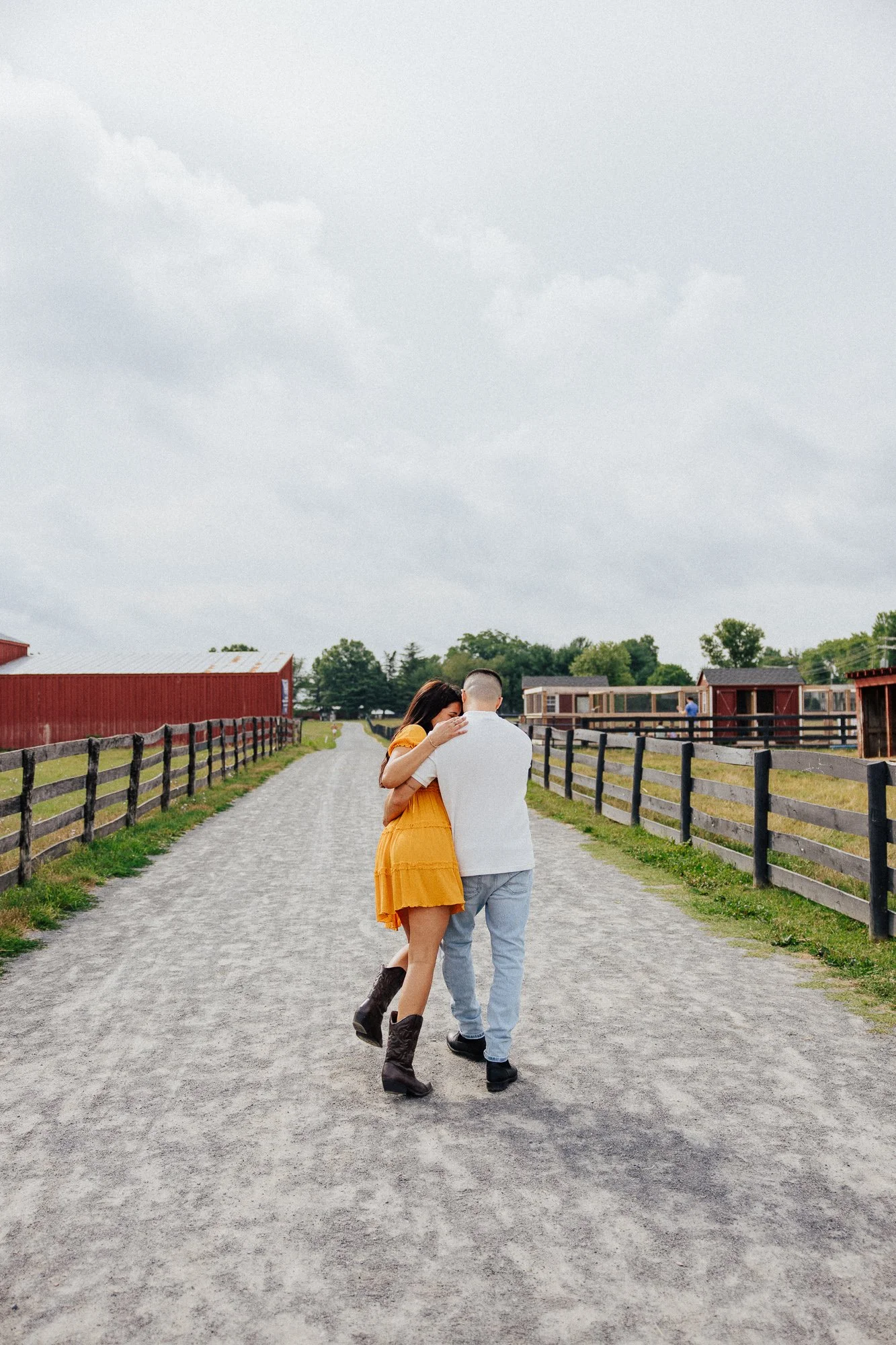 A man and woman walking together on a gravel path, holding each other, with a cloudy sky and a rural setting with barns and trees in the background.