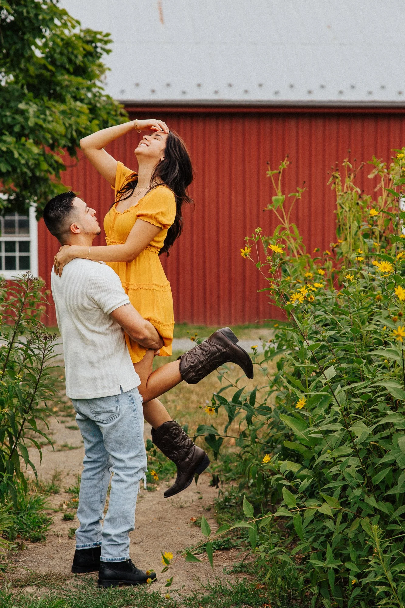 A man is holding a woman in his arms outdoors near yellow flowers with a red barn in the background. The woman is wearing a yellow dress and has her hand on her forehead, smiling with her eyes closed. The man is wearing a light-colored polo and gray 