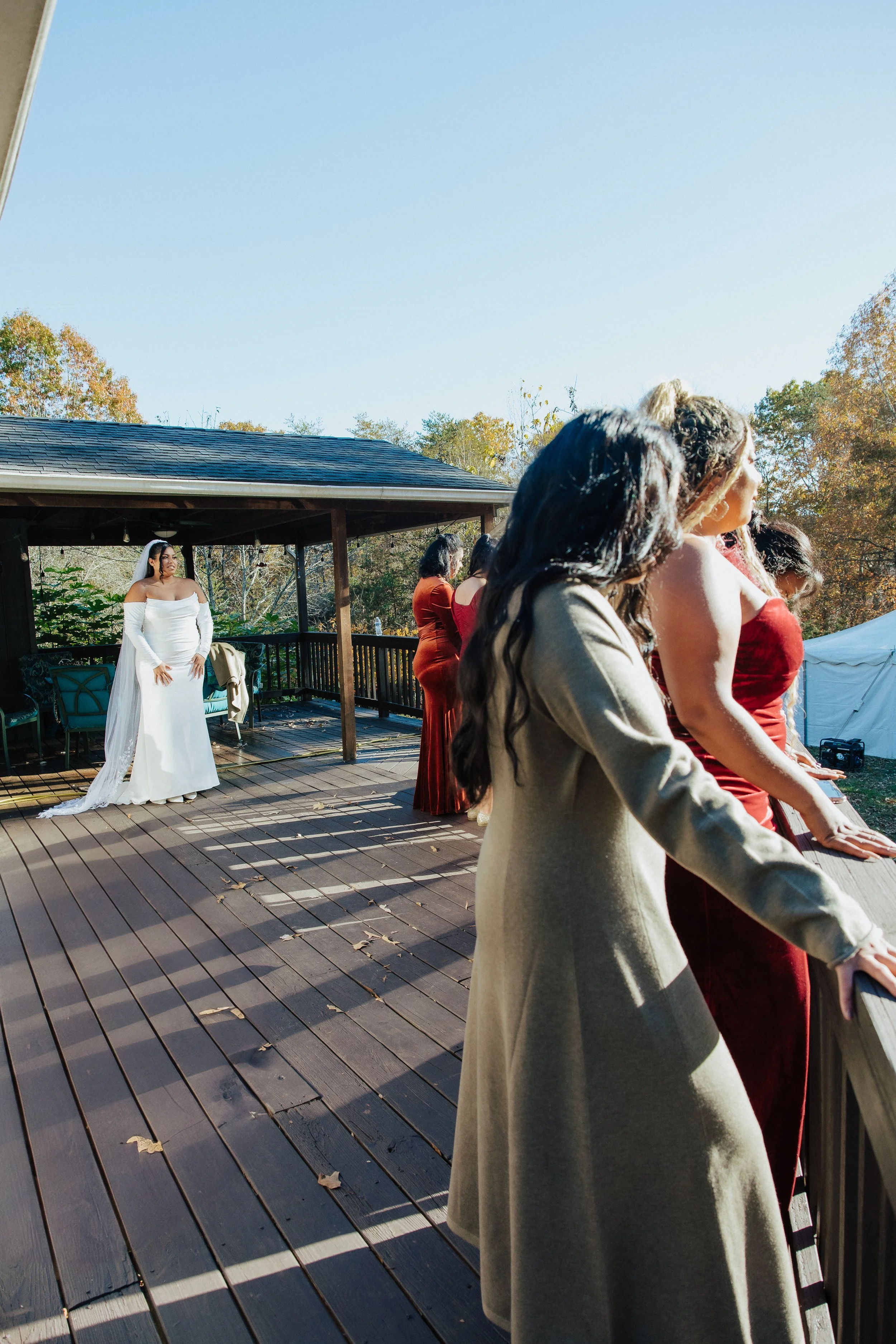 Women in formal dresses and a bride in a white wedding gown are gathered outdoors on a wooden deck, with trees in the background and a blue sky overhead.