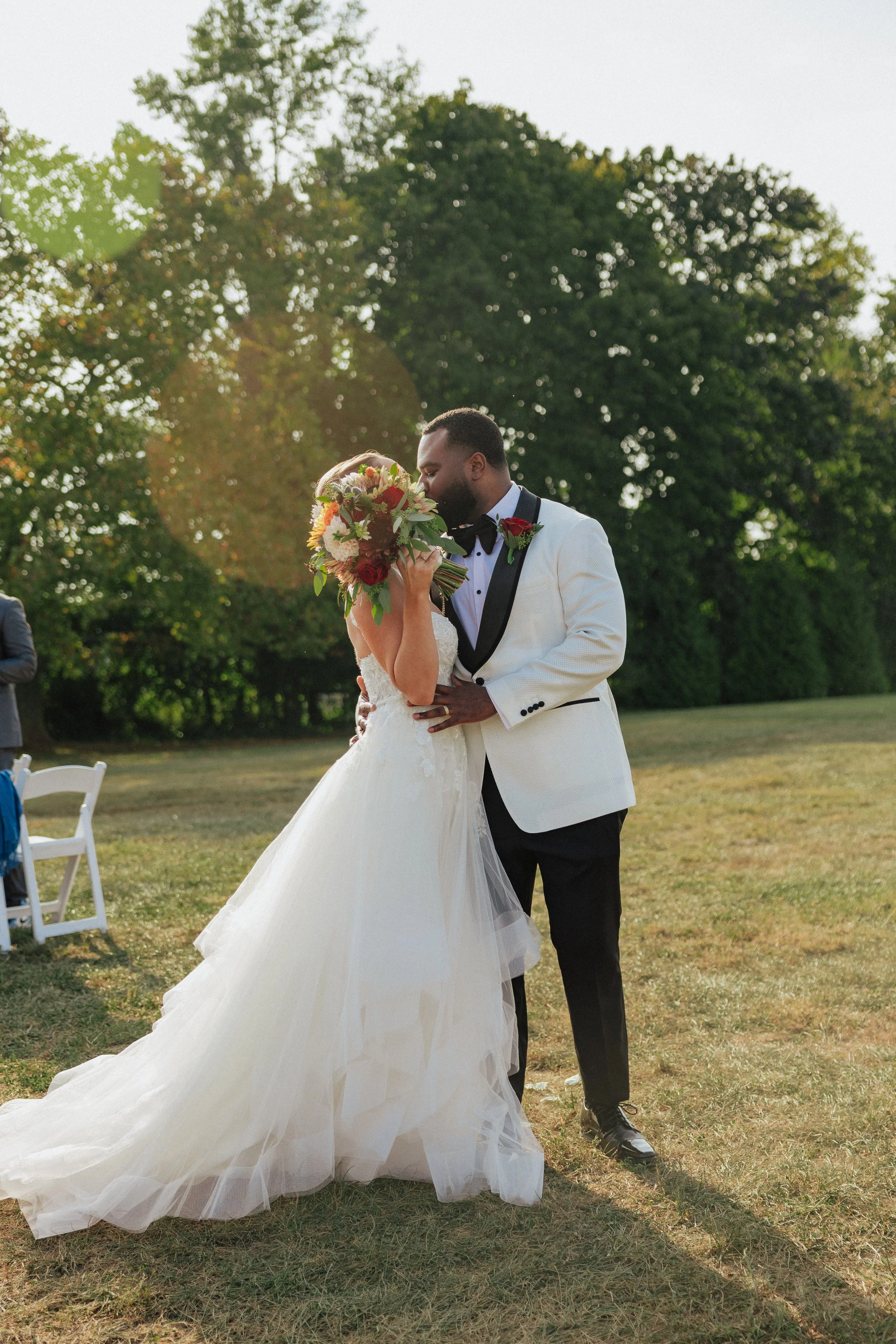 Bride and groom sharing a kiss outdoors during a wedding ceremony, with trees in the background and sunlight filtering through.