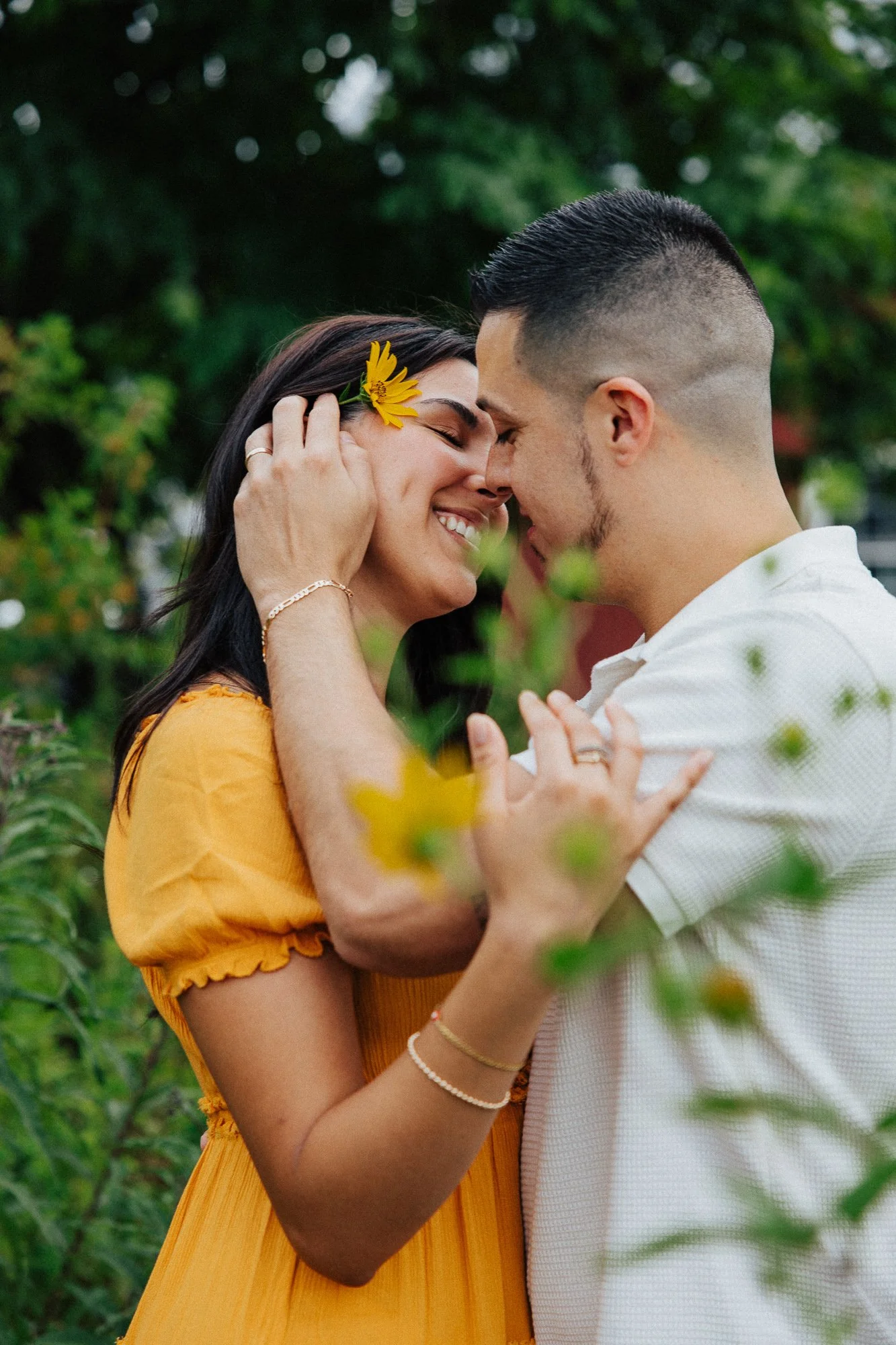 A couple touching foreheads and smiling outdoors, with a woman wearing a yellow dress and a yellow flower in her hair, and a man in a white shirt, surrounded by green foliage.