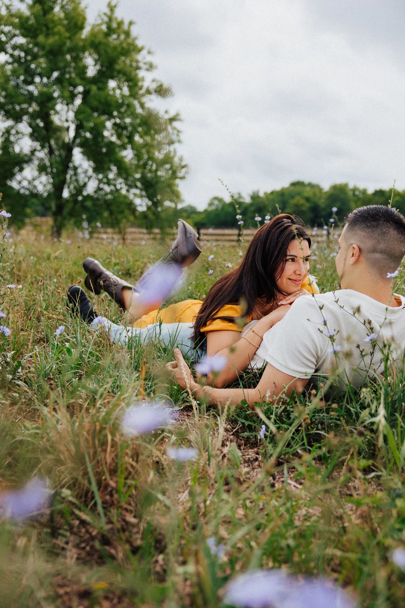 A couple lying on the grass in a field with flowers, facing each other and smiling.